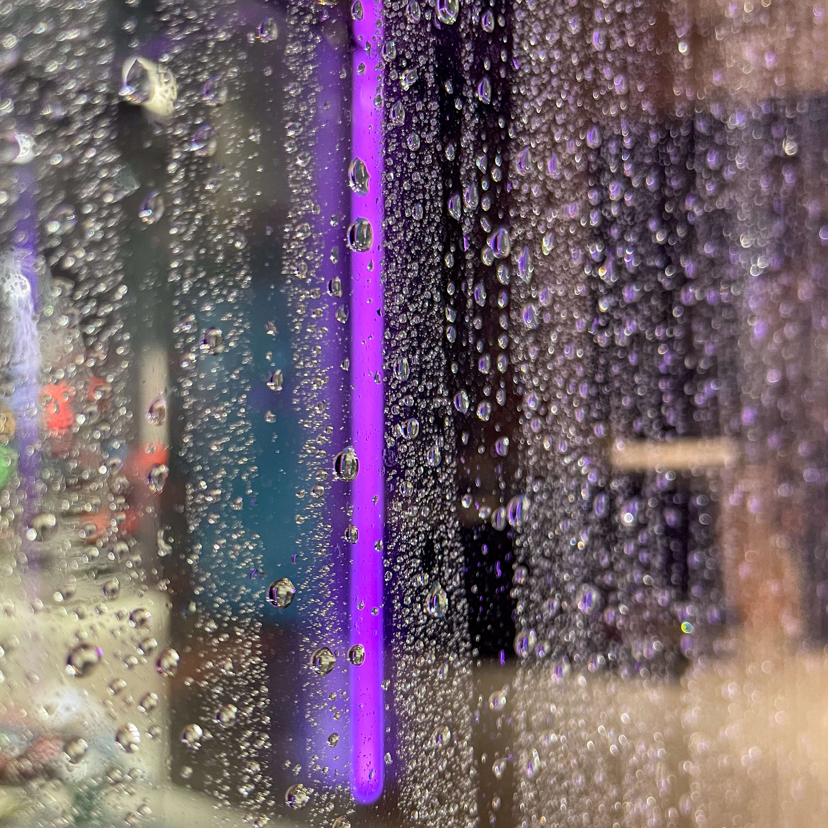 A close-up of a glass surface covered in water droplets. A purple light is shining through the droplets. The image is out of focus.