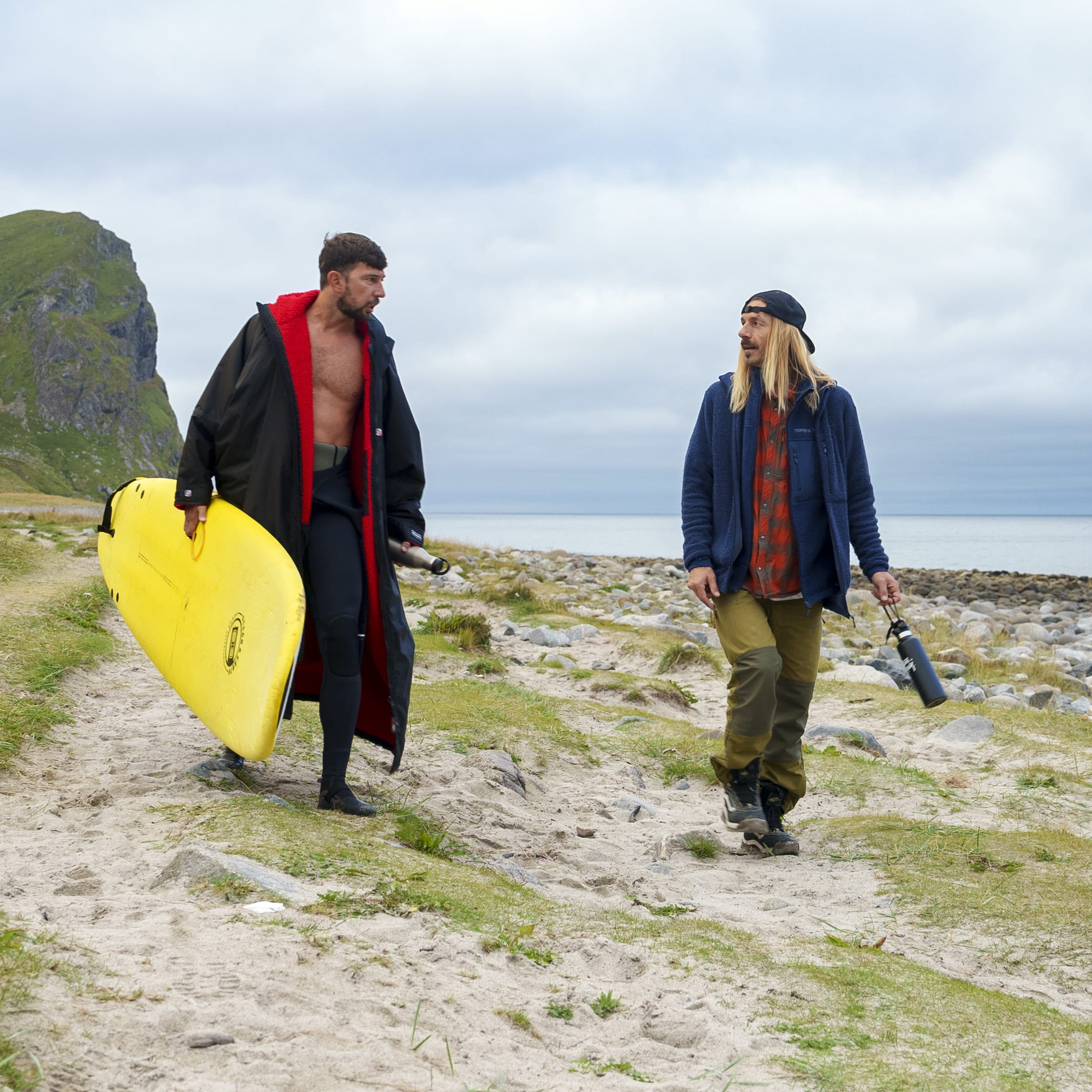 Two men walking on a sandy beach. One man is carrying a yellow surfboard and wearing a Norrøna waterproof jacket. The other man is carrying a water bottle.