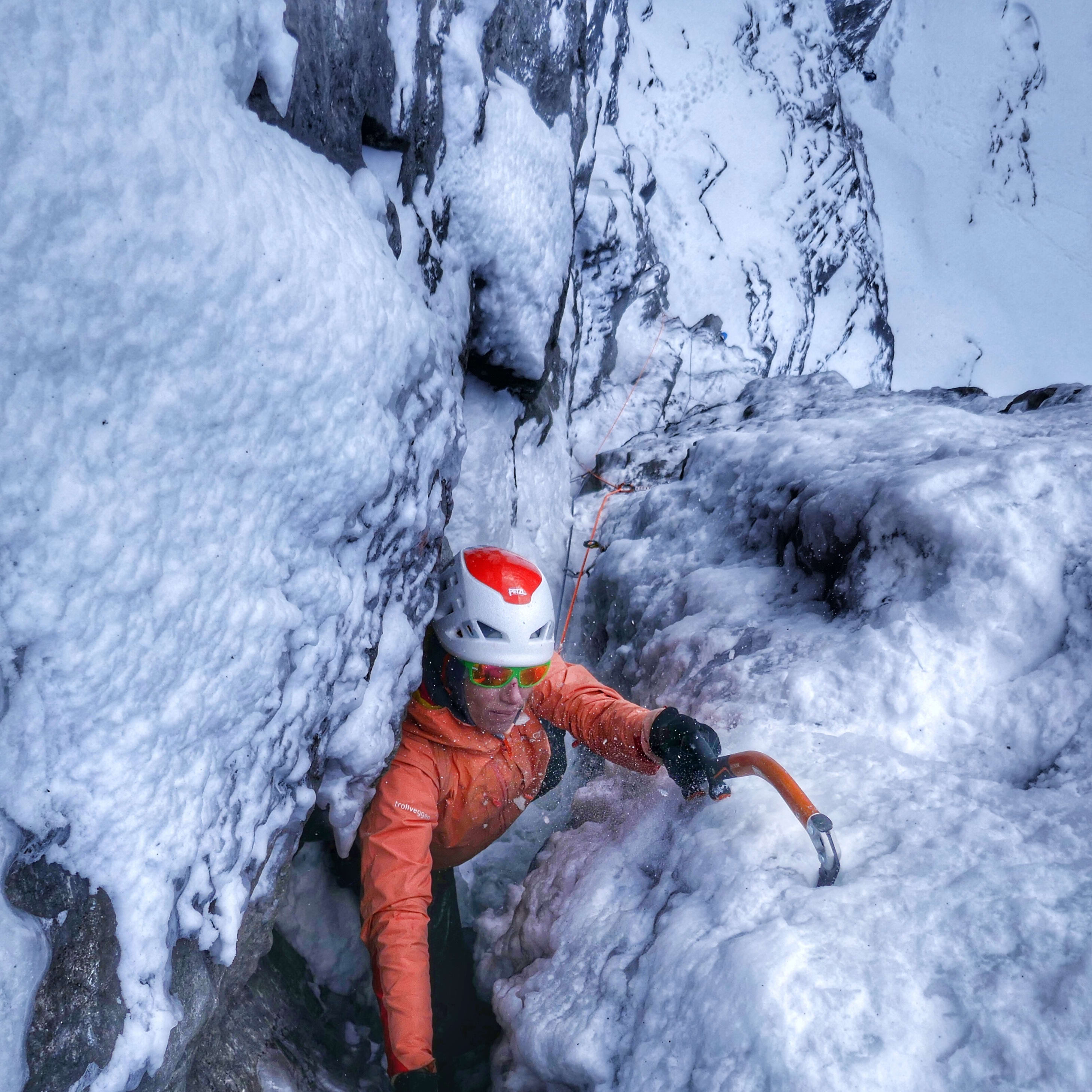Ice climber in orange Norrøna jacket and helmet, using ice axes to climb a narrow, icy chute.