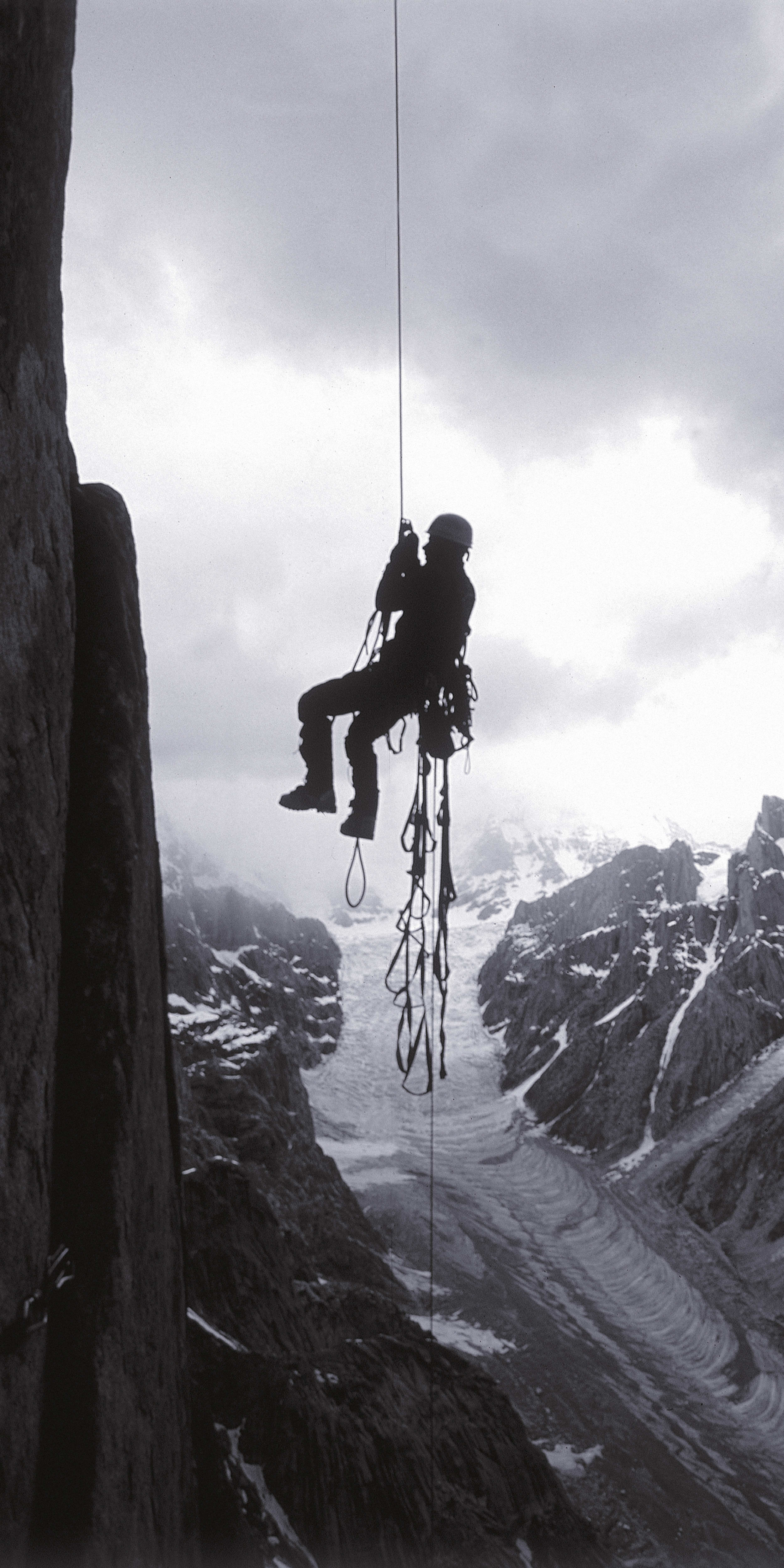 A silhouetted climber rappels down a steep rock face with a glacier in the background.