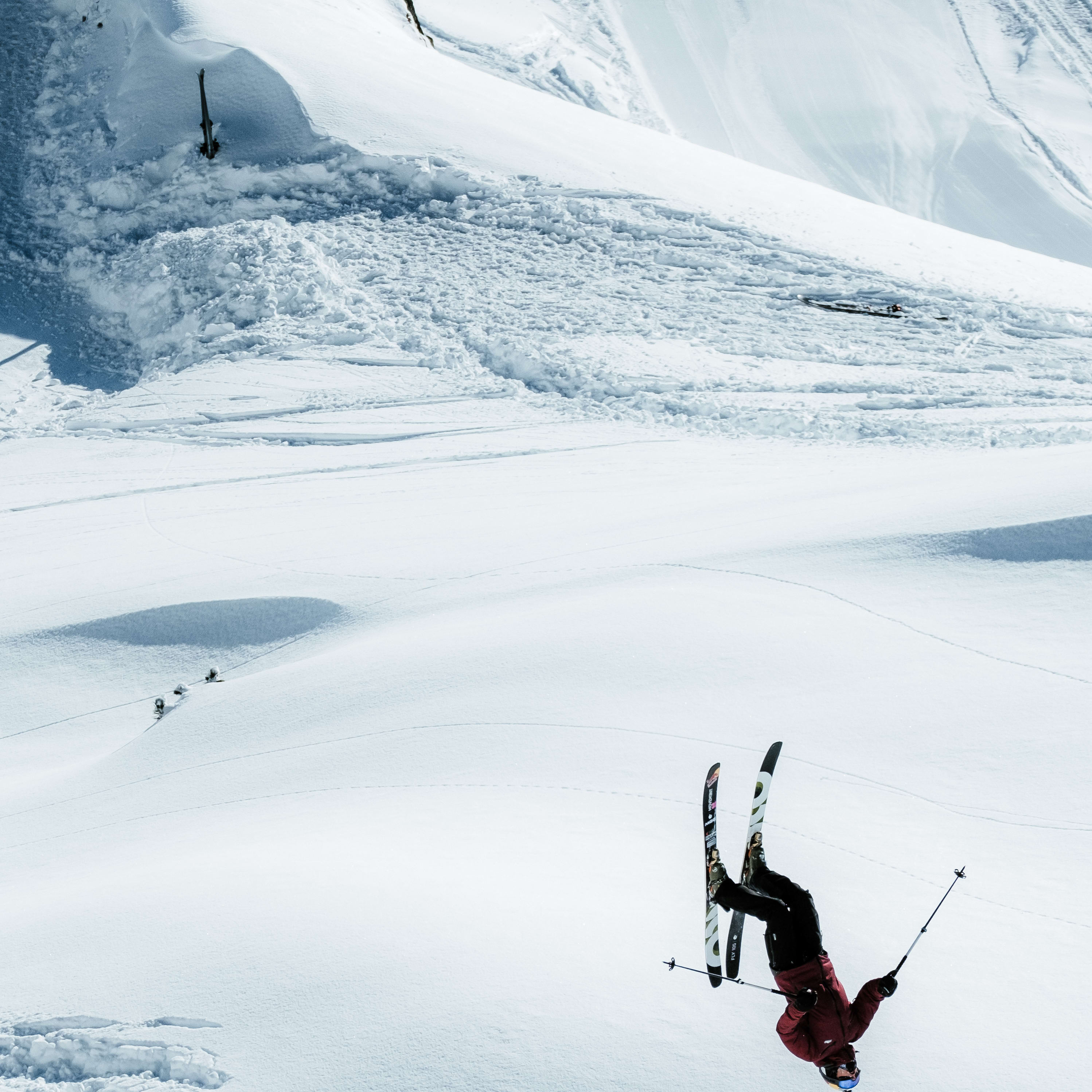 A skier in mid-air, performing a trick in a snowy mountain landscape. The skier wears a red Norrøna jacket.