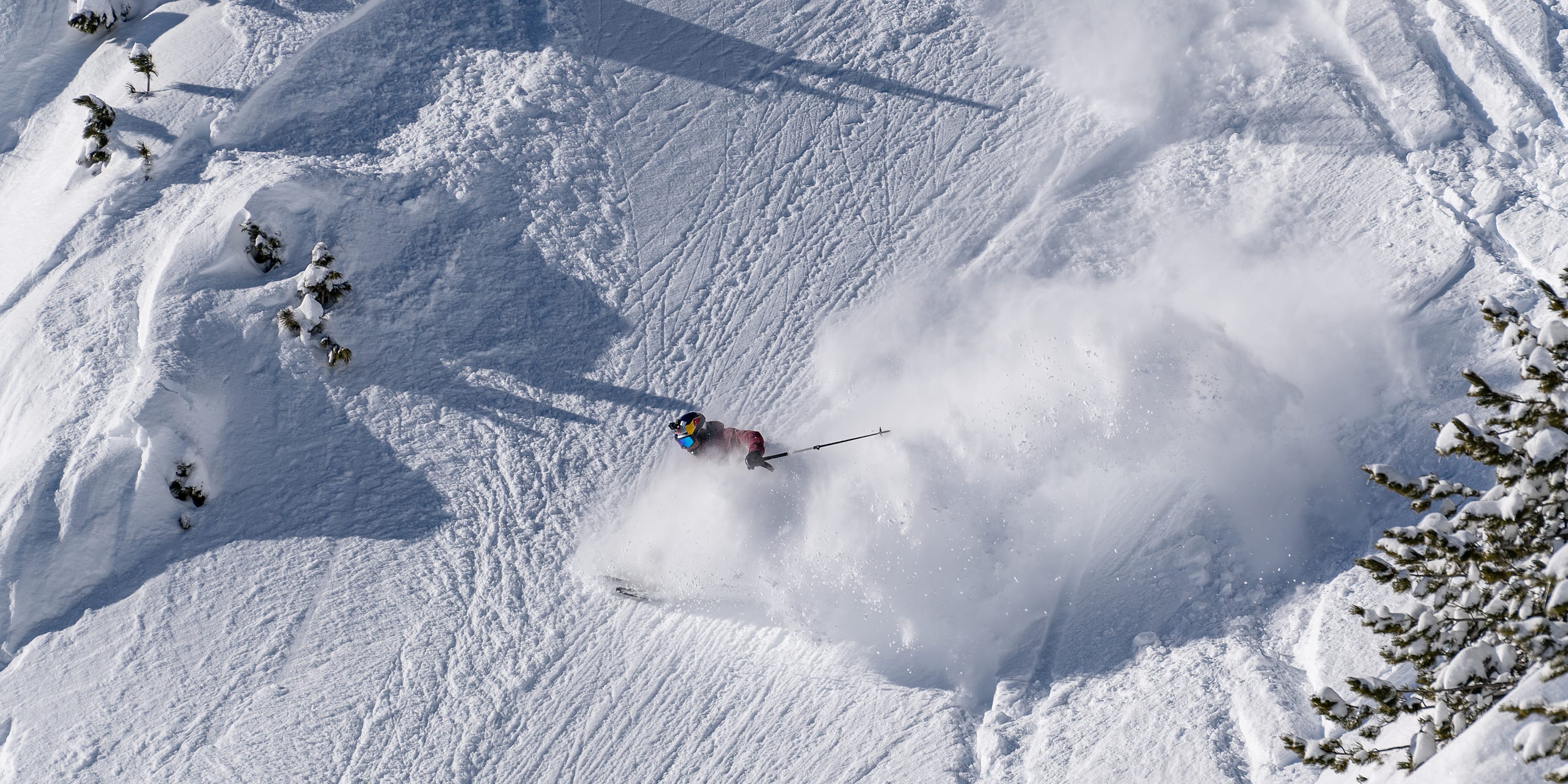 Aerial view of a skier in red Norrøna jacket, amidst snow spray, skiing down a steep, snowy mountain slope.