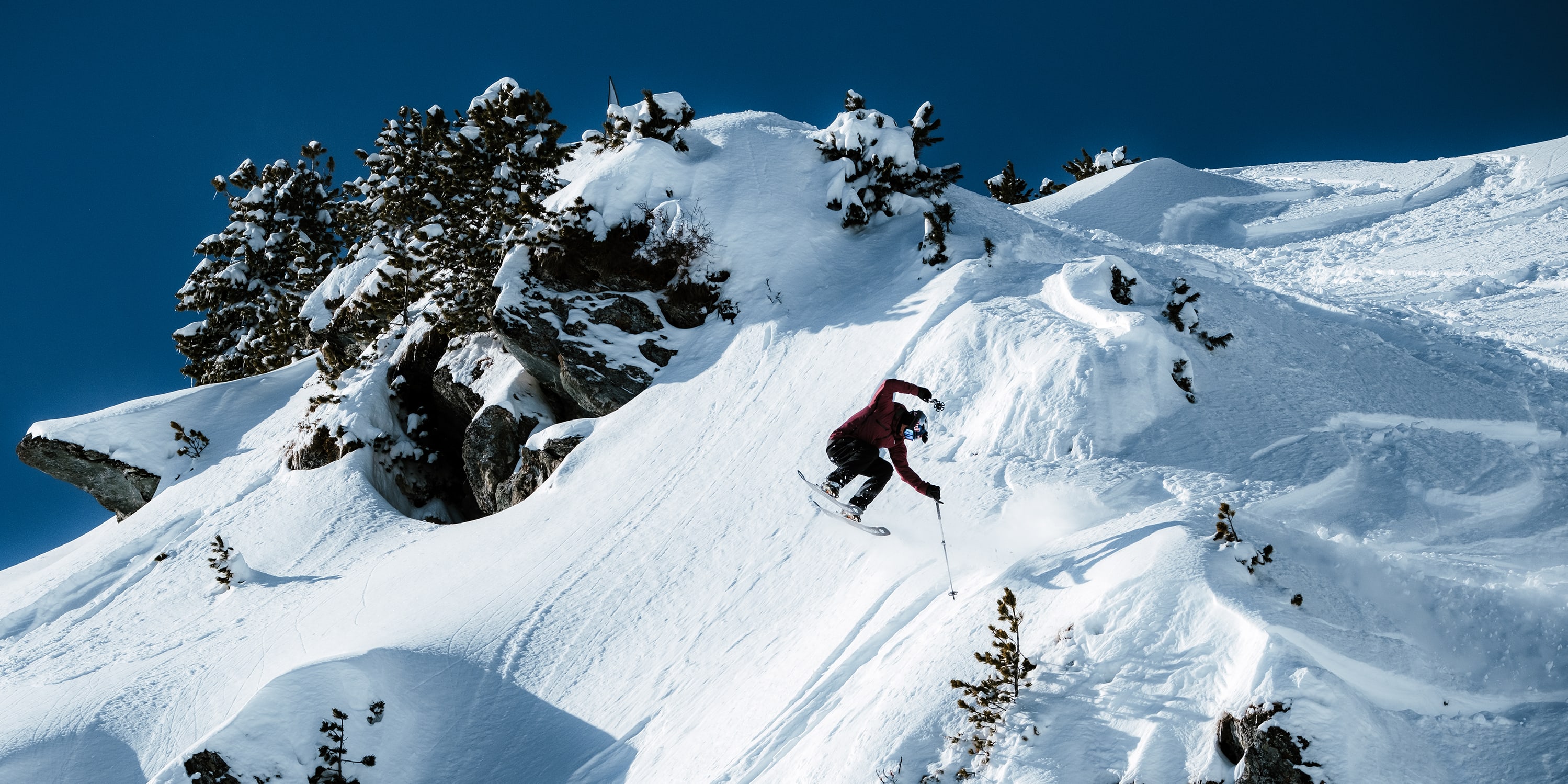 A skier wearing a Norrøna jacket jumps on a snowy mountain under a bright blue sky.
