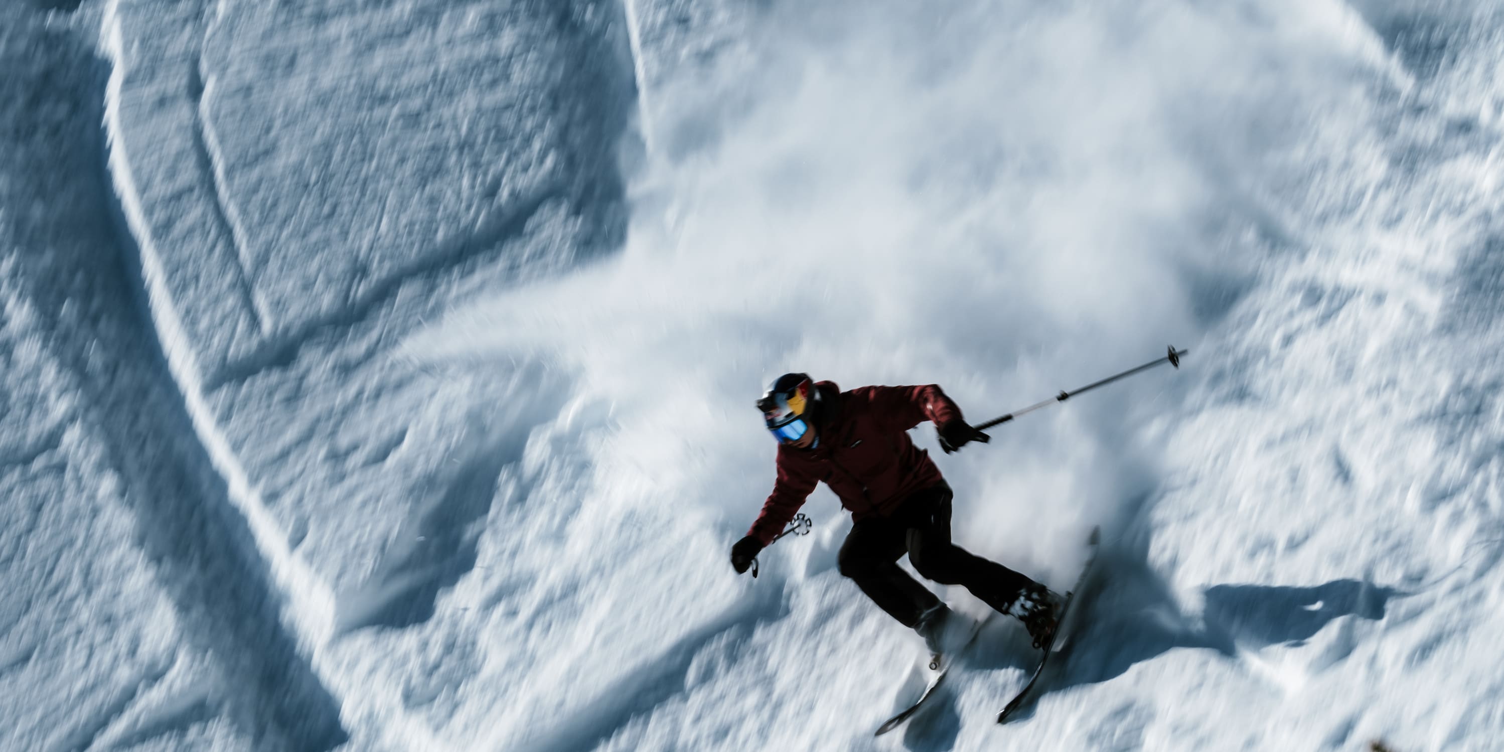 A skier in a red Norrøna jacket carving through fresh powder. The action is captured from above creating motion.