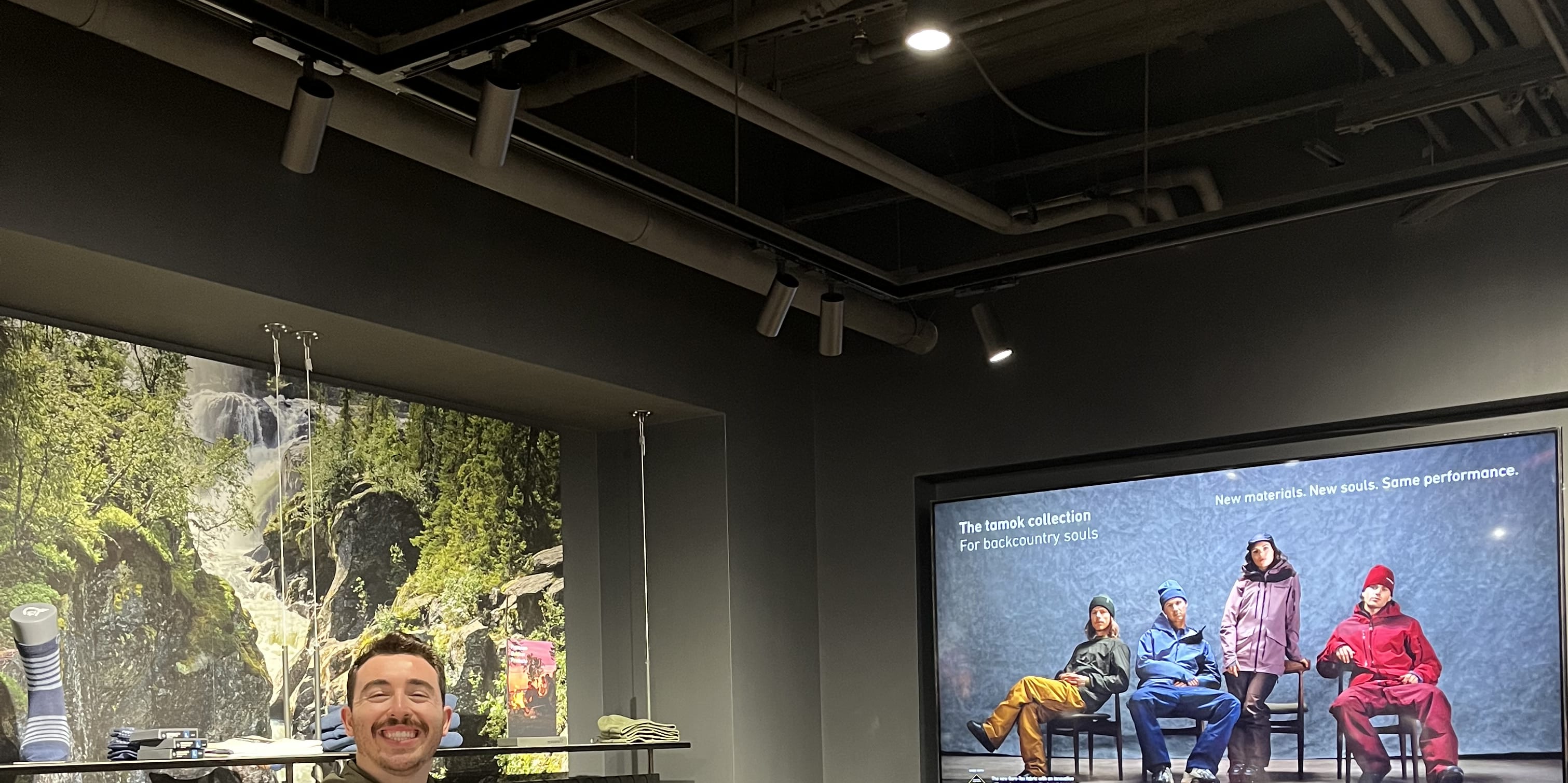 A man in a Norrøna sweater stands smiling in a store with clothing racks and a screen showing a model wearing Norrøna outdoor apparel. 
