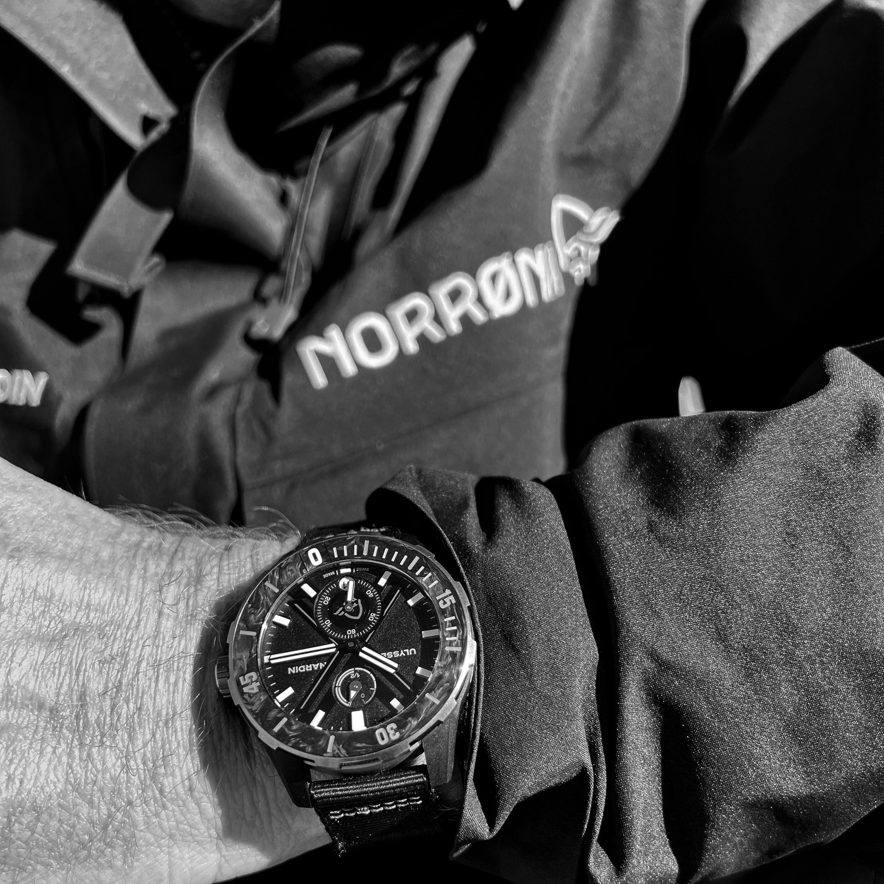 A black and white photo of a wrist wearing a black Norrøna jacket and a black wristwatch. 
