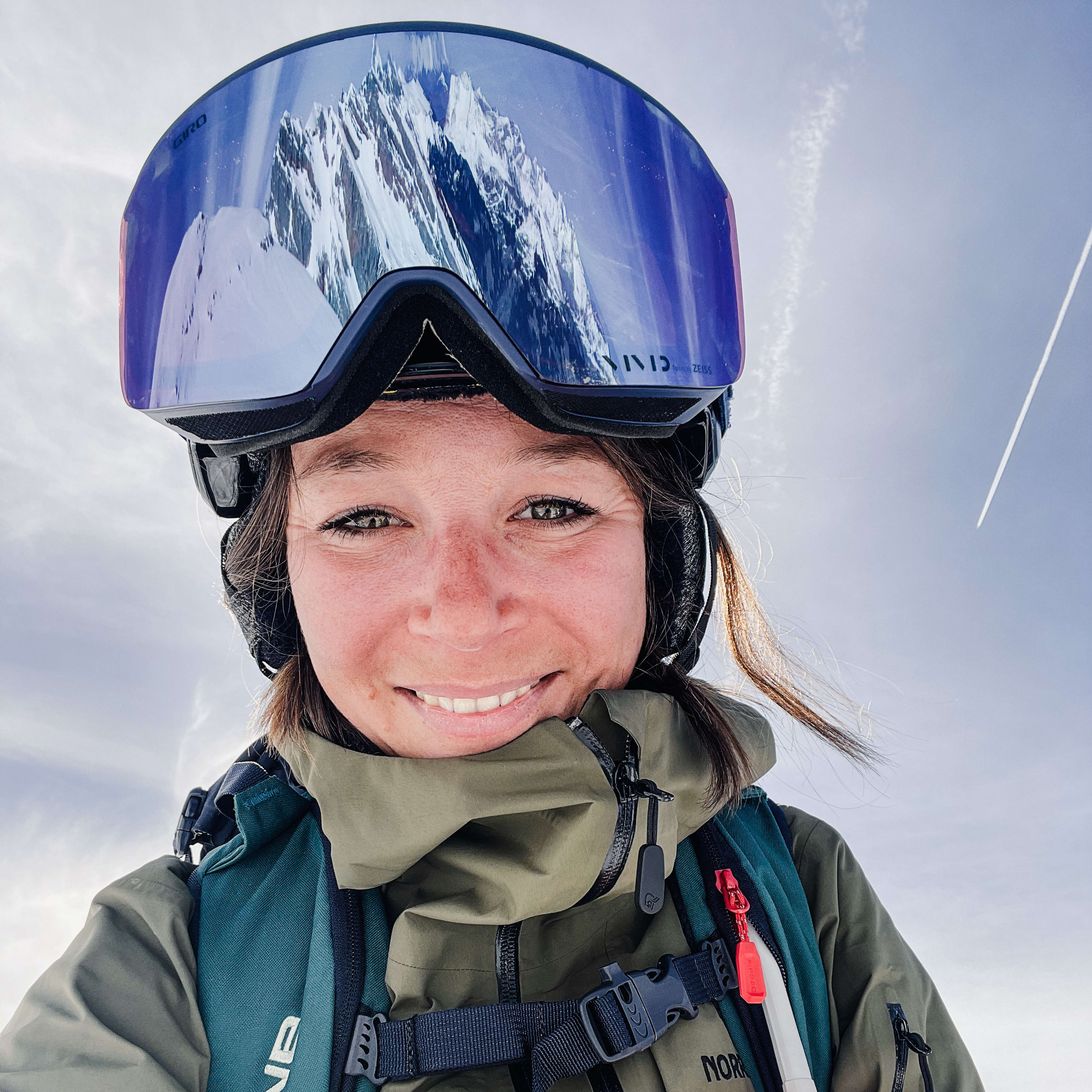 Close-up of a smiling woman in snow gear, including a Norrøna jacket and Giro goggles reflecting a mountain. Sky background with a contrail.
