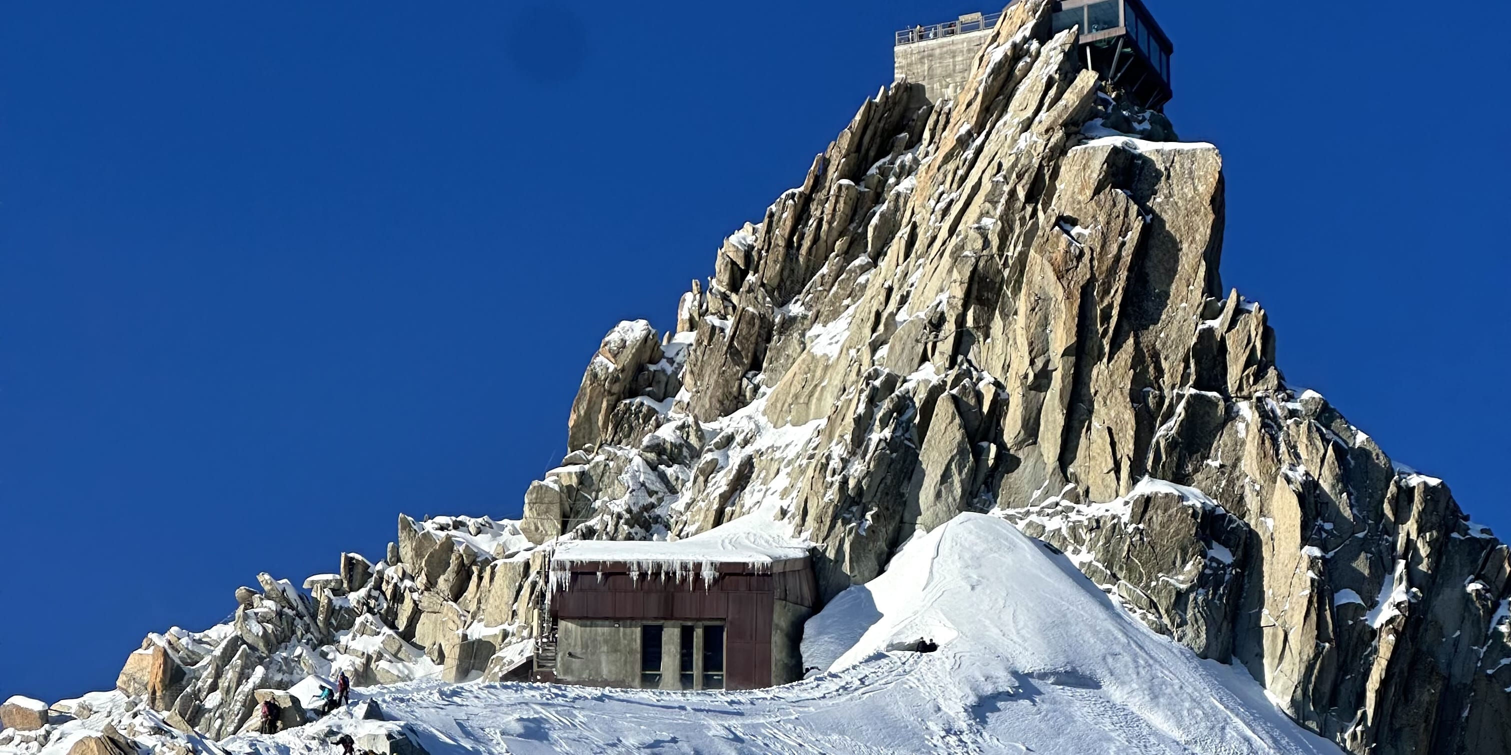 Aiguille du Midi mountain view, climbers ascending a snowy slope, building with a tower on top, clear blue sky. Some wearing Norrøna clothing.