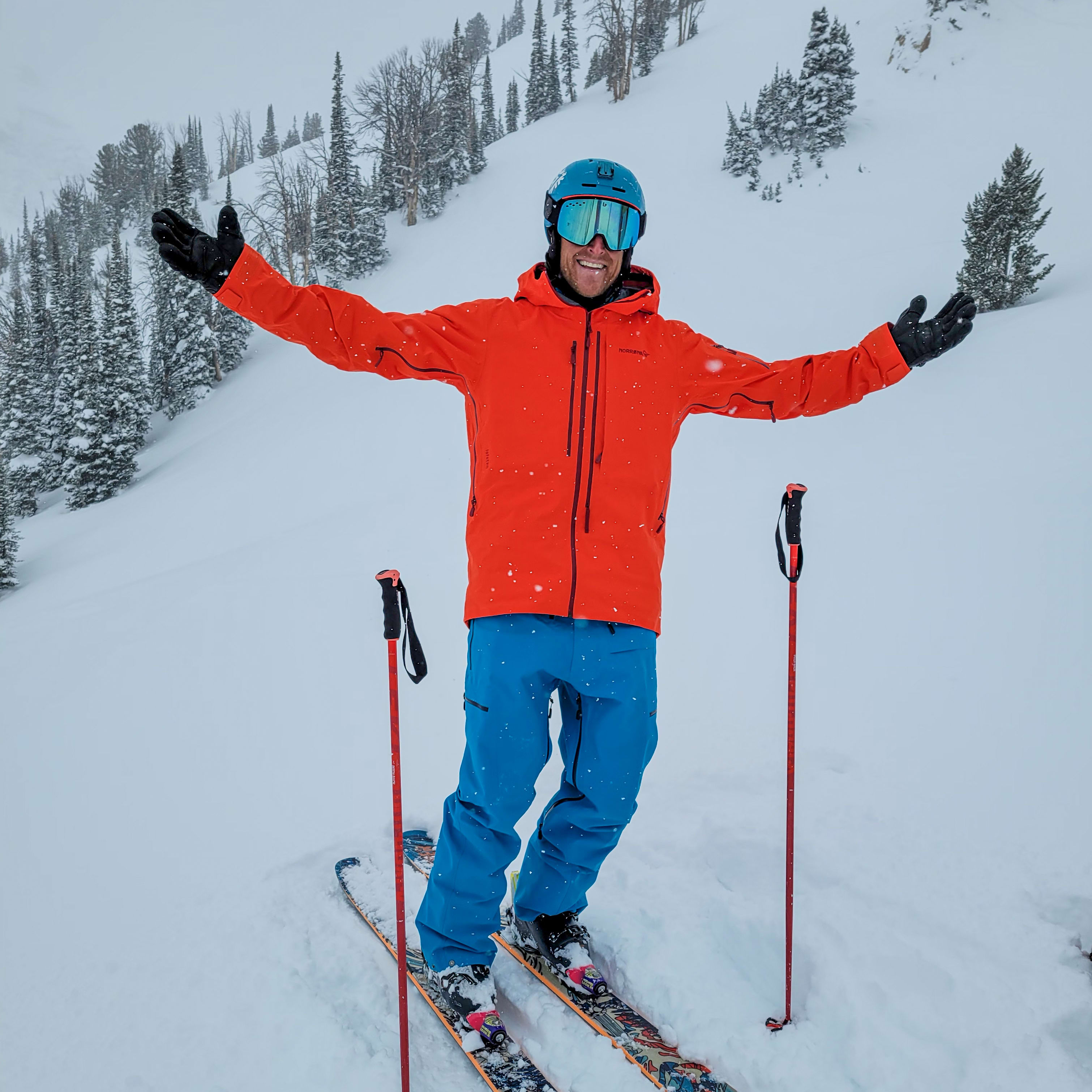 A skier with arms outstretched on a snowy slope. He wears a Norrøna jacket and pants, helmet, and goggles. Ski poles are beside him.