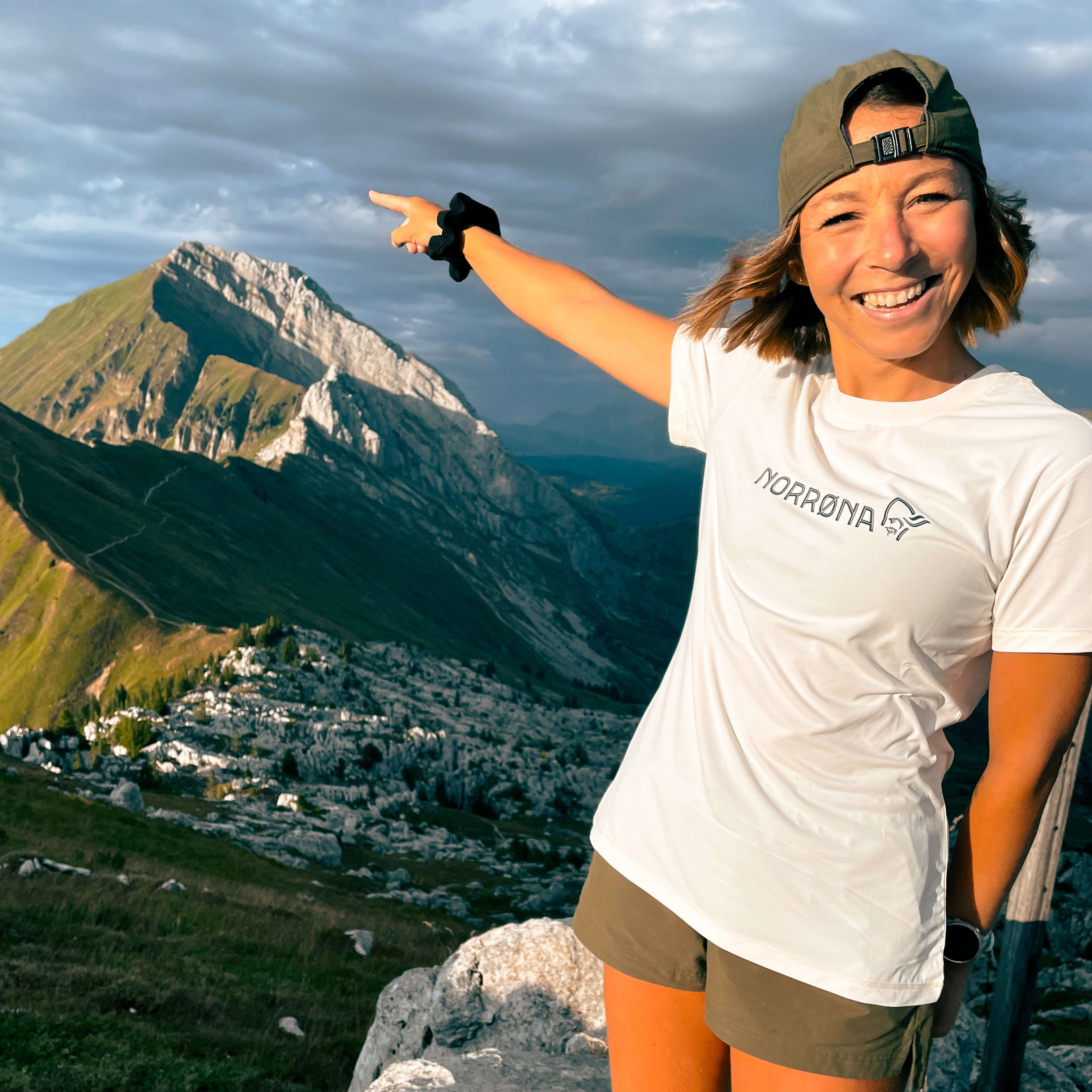 A smiling woman wearing a Norrøna t-shirt and cap points to a mountain range in the background.