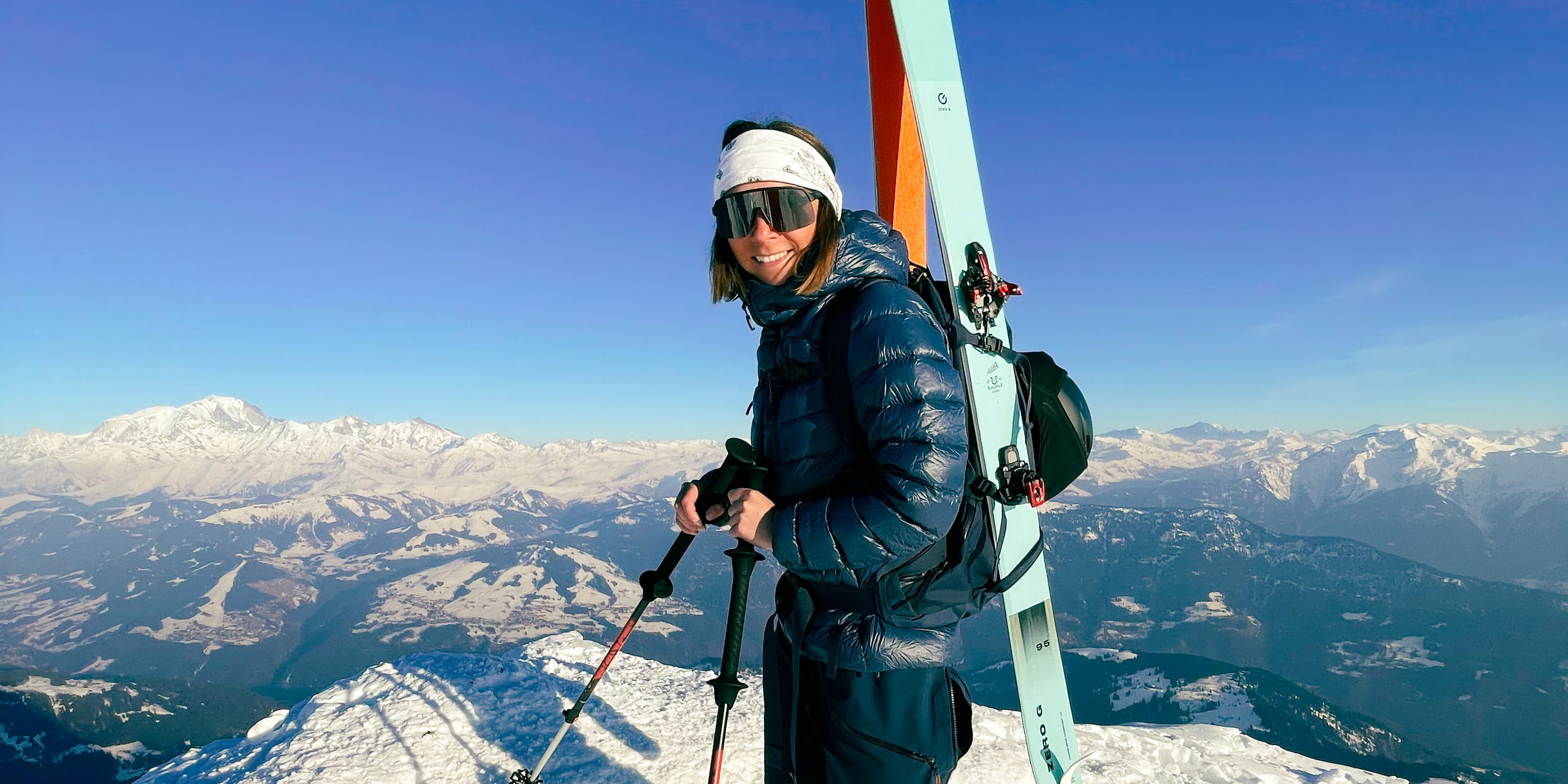 A woman on a snowy mountaintop wears Norrøna ski apparel and carries skis and poles with snow-capped mountains in the background.