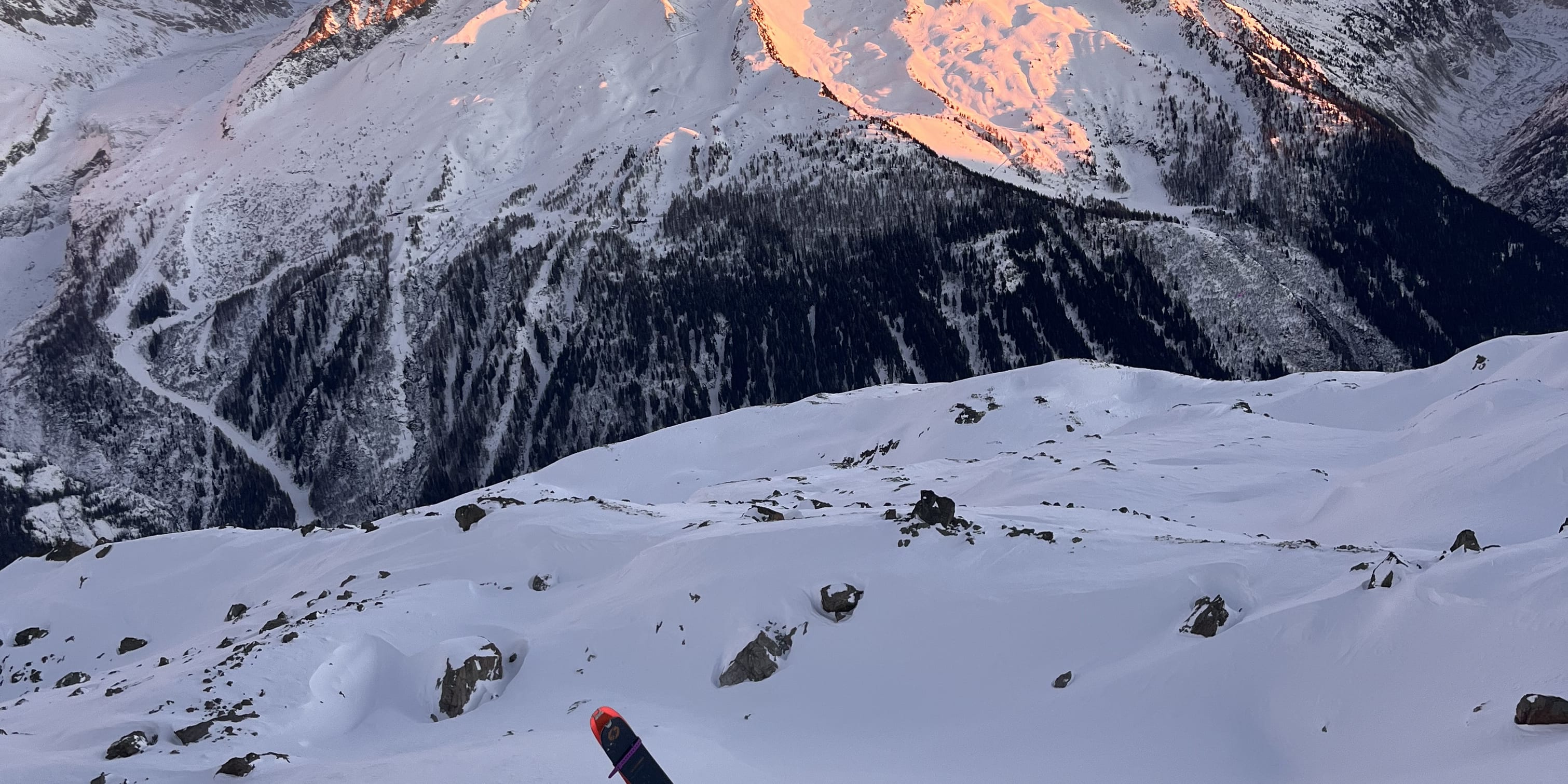 A person wearing Norrøna gear and skis walks up a snowy mountain slope with a large mountain range in the background. 
