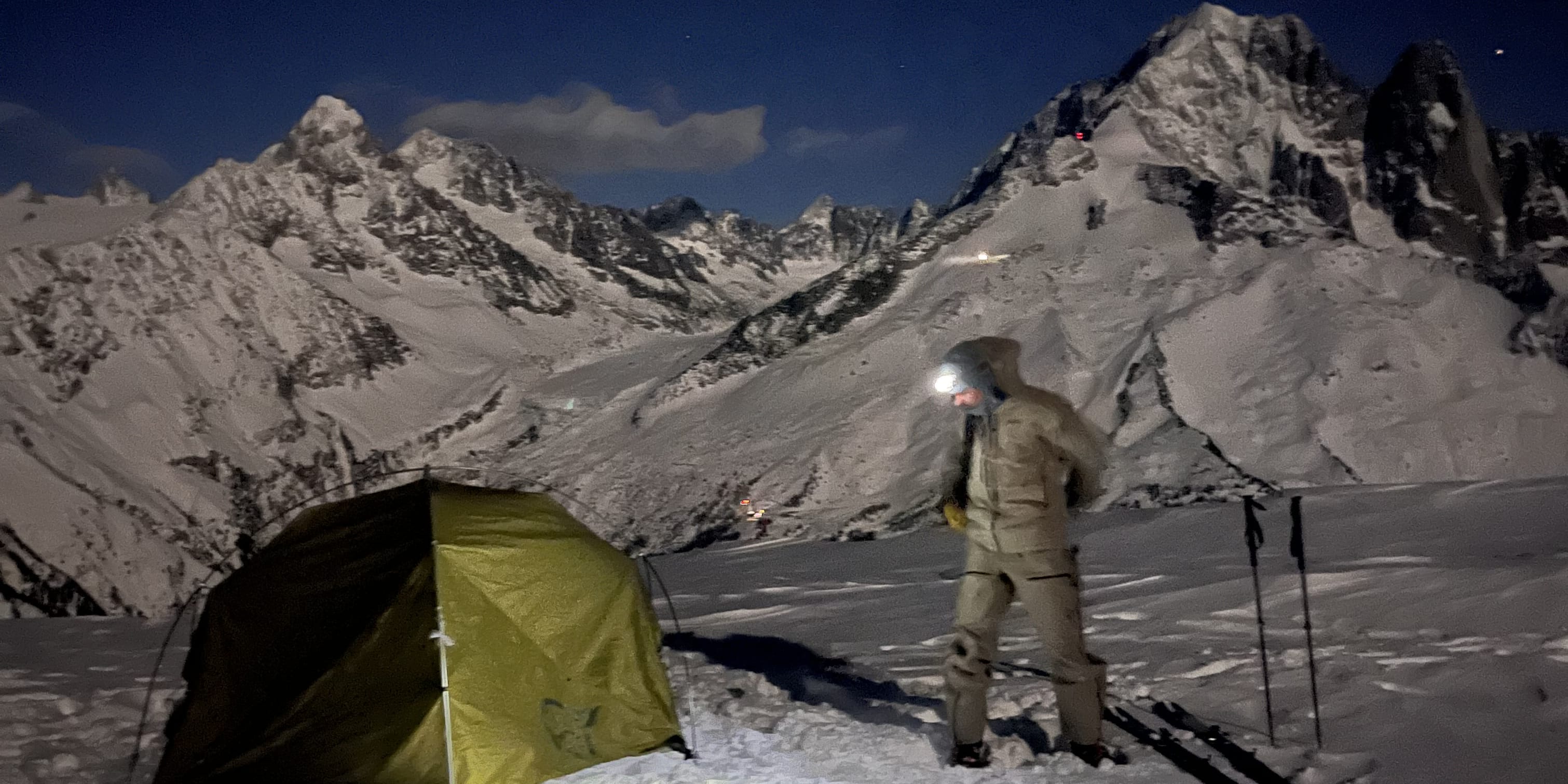 Person in Norrøna clothing standing in snow with a headlamp on, next to a green tent and snowy mountains in the background. 
