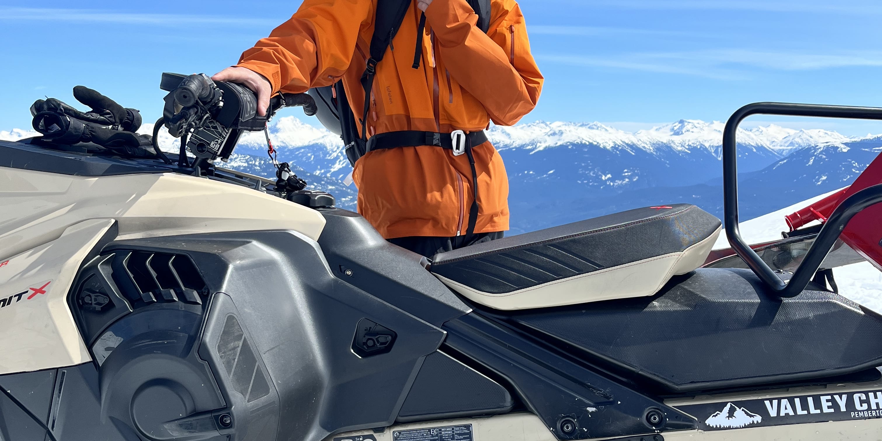 A snowmobiler in a helmet and orange Norrøna jacket stands on their snowmobile in a snowy mountain landscape under a blue sky.
