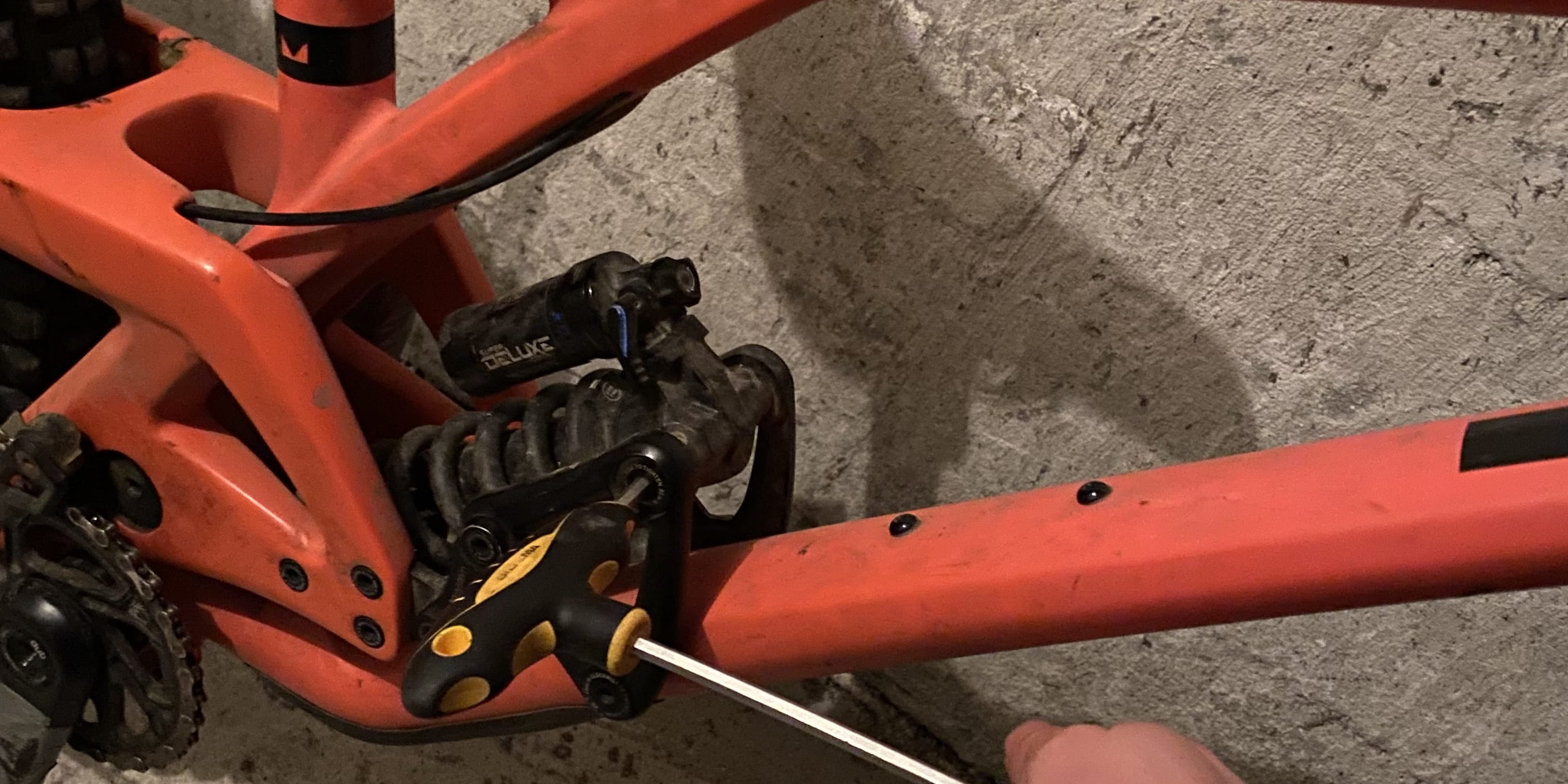 A close-up of a red mountain bike with a black shock absorber. A person is using an Allen key to tighten a bolt on the bike frame.