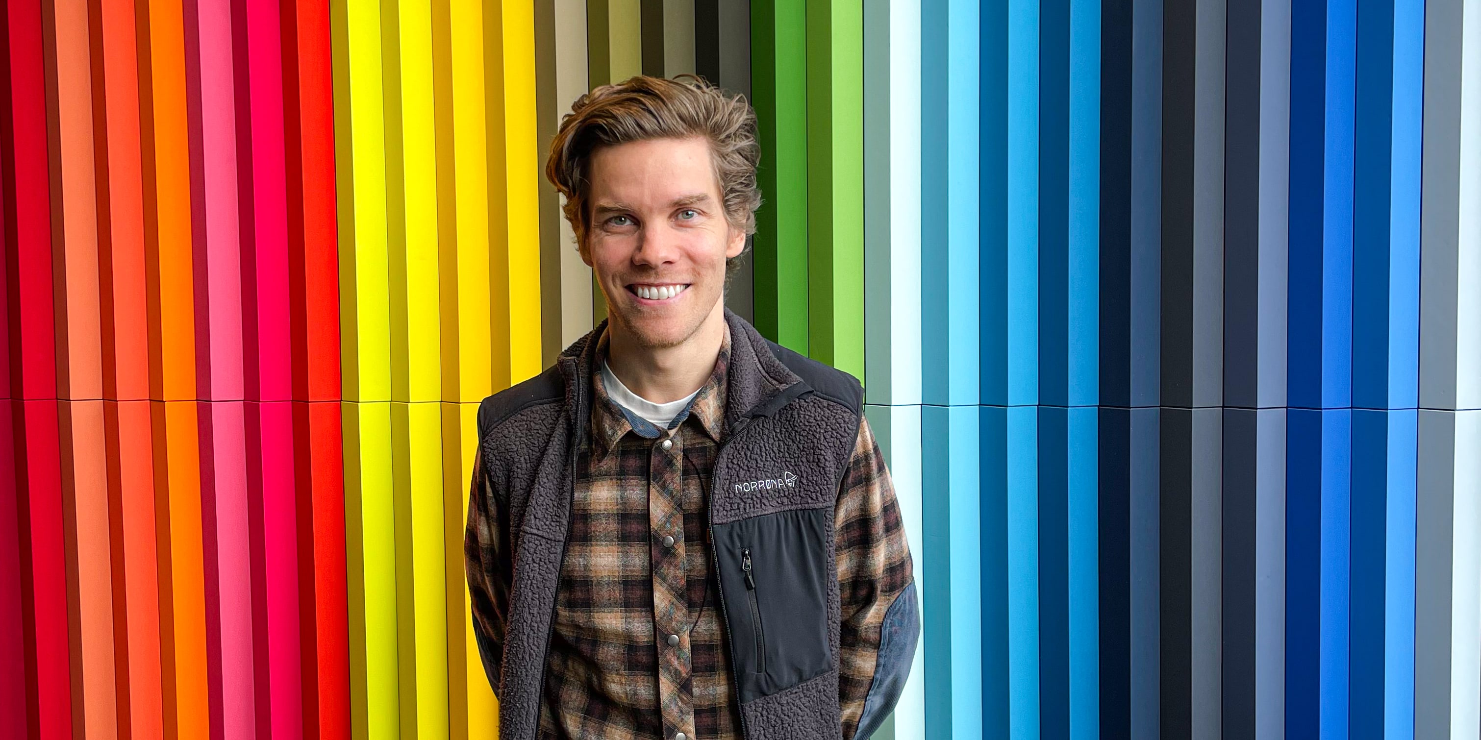 A man in a plaid shirt and black vest stands in front of a wall of vertical colorful panels. The wall has the words "Norrøna House" in white lettering.  The man is smiling at the camera.