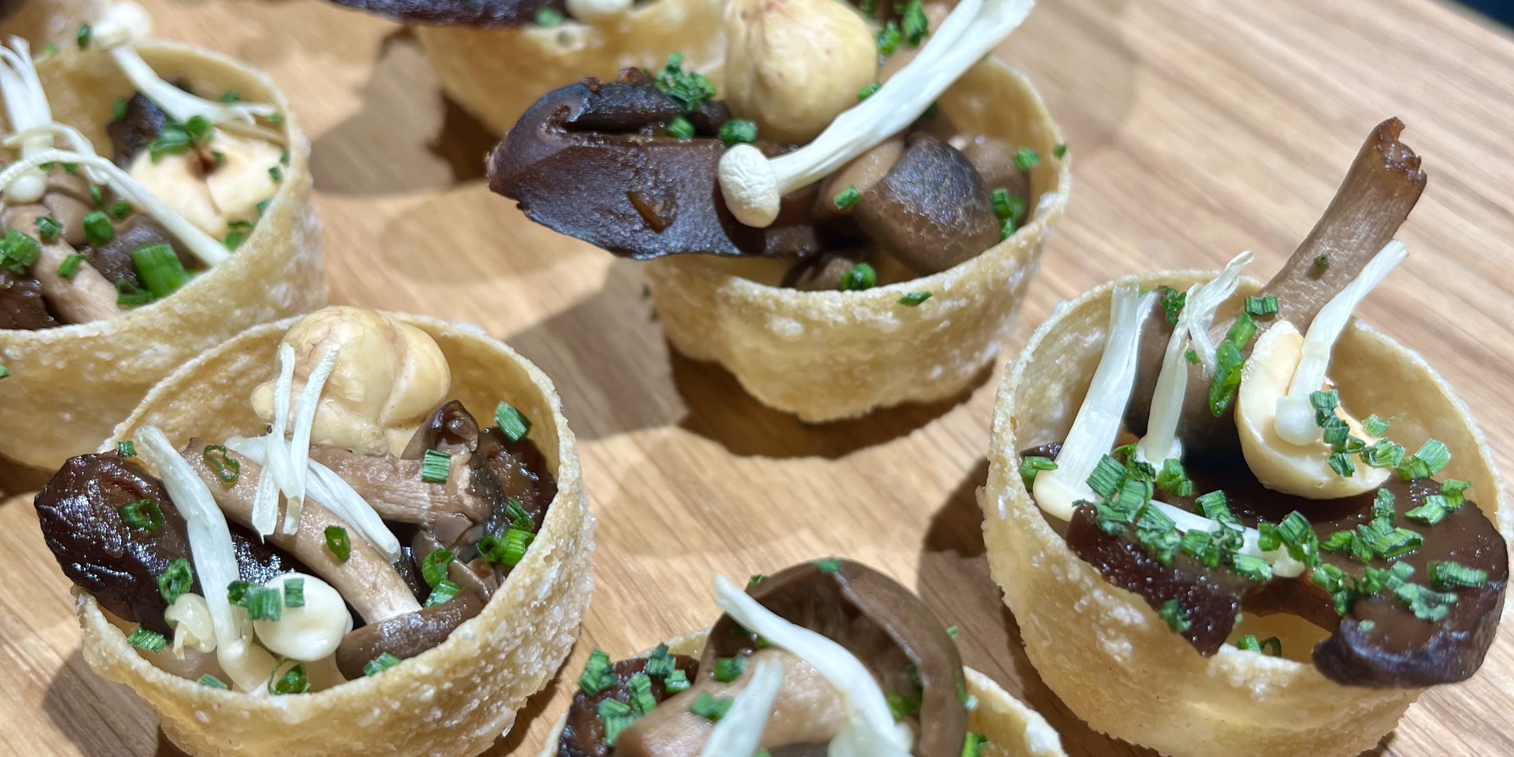 Closeup of small edible bowls filled with mushrooms, hazelnuts, and chives on a wooden surface.