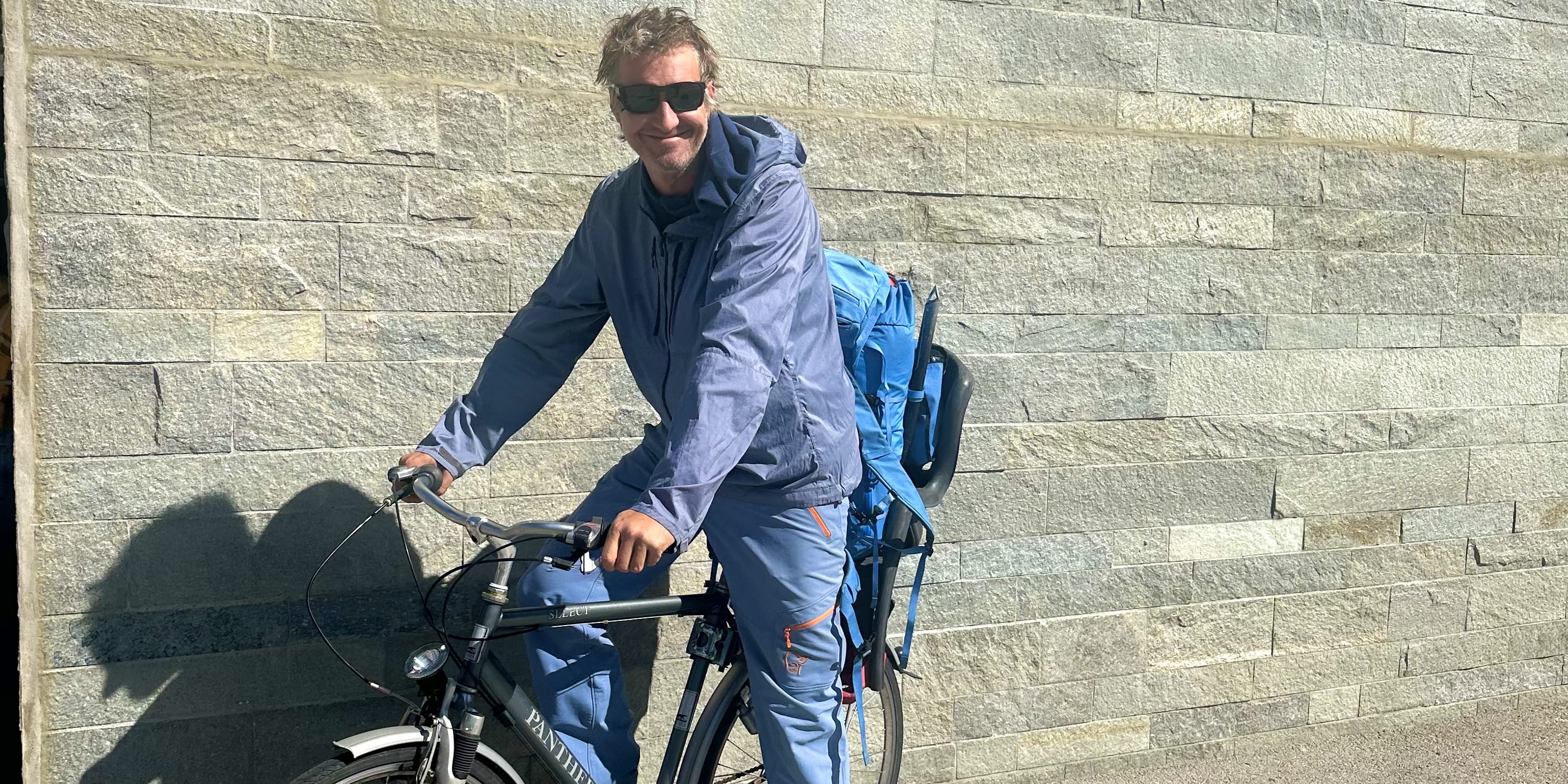 Man on a "Panther" bicycle with a child seat. He wears Norrøna jacket & pants, and orange boots. Stone wall background.