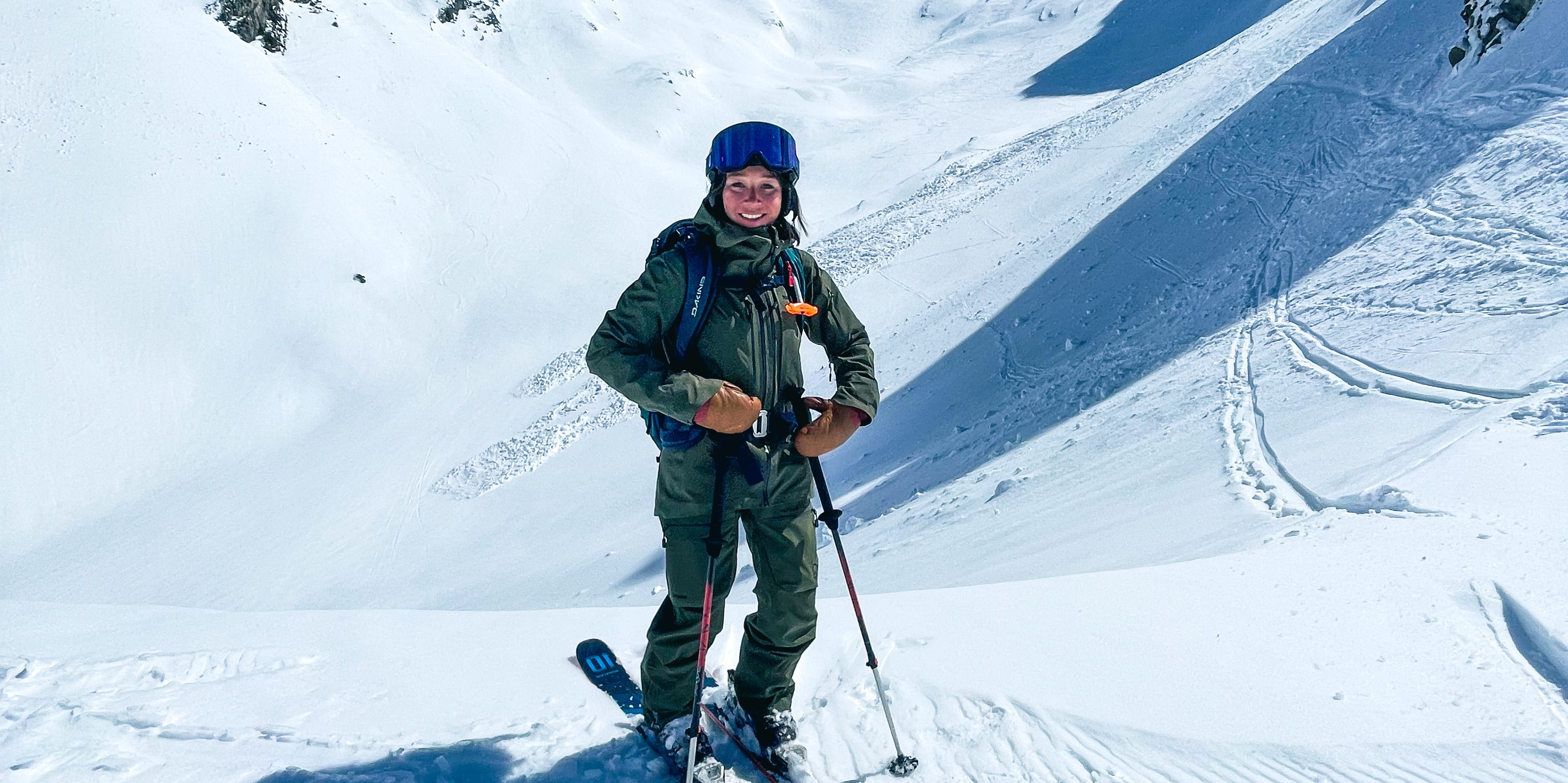 A skier wearing a blue helmet and a green Norrøna suit stands on snow-covered mountain with tall, rocky peaks under a clear blue sky.