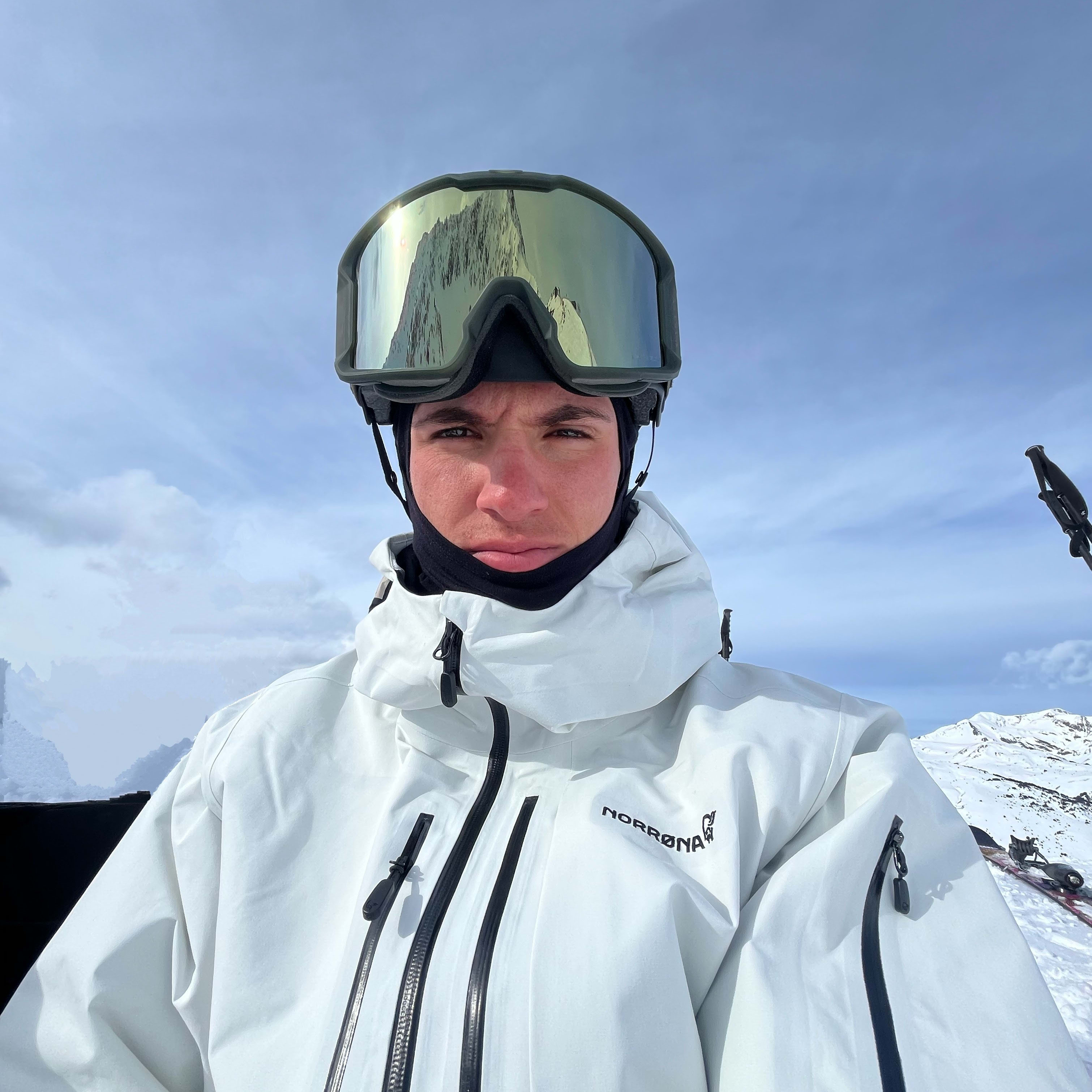 A close-up of a skier wearing a Norrøna jacket and goggles, reflecting a snowy mountain. The sky is partly cloudy.