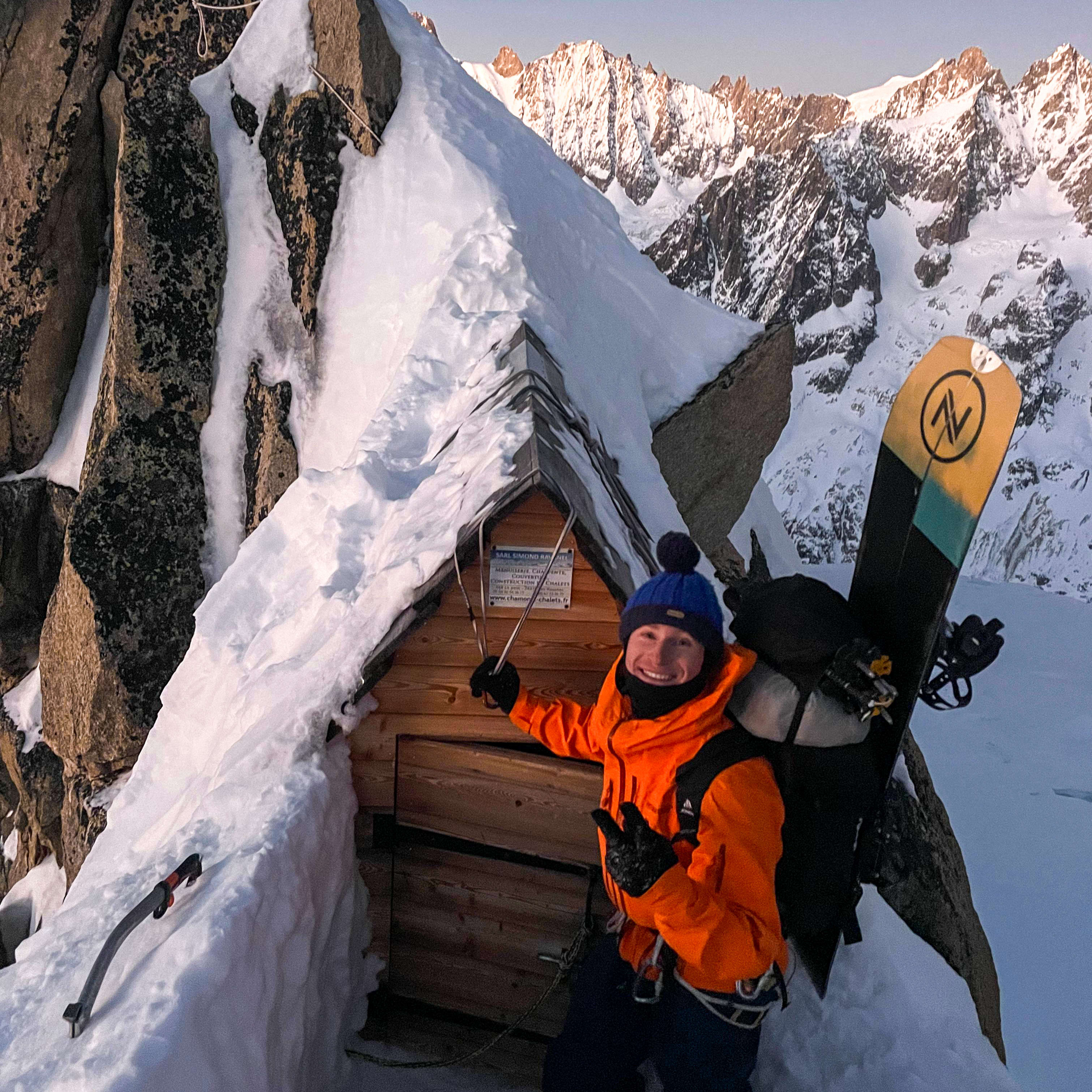 A man wearing an orange Norrøna jacket and a blue beanie smiles near a small wooden cabin, standing on a snowy mountaintop.