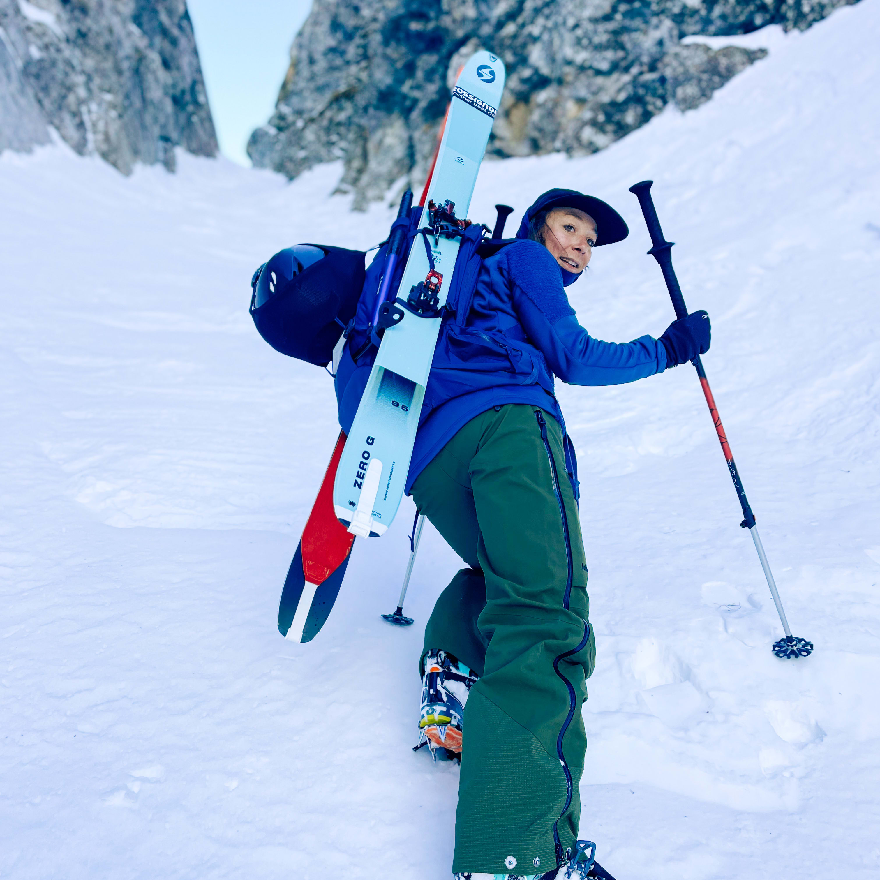 A person in Norrøna jacket & pants, climbing snowy terrain with skis and poles. Mountain backdrop.