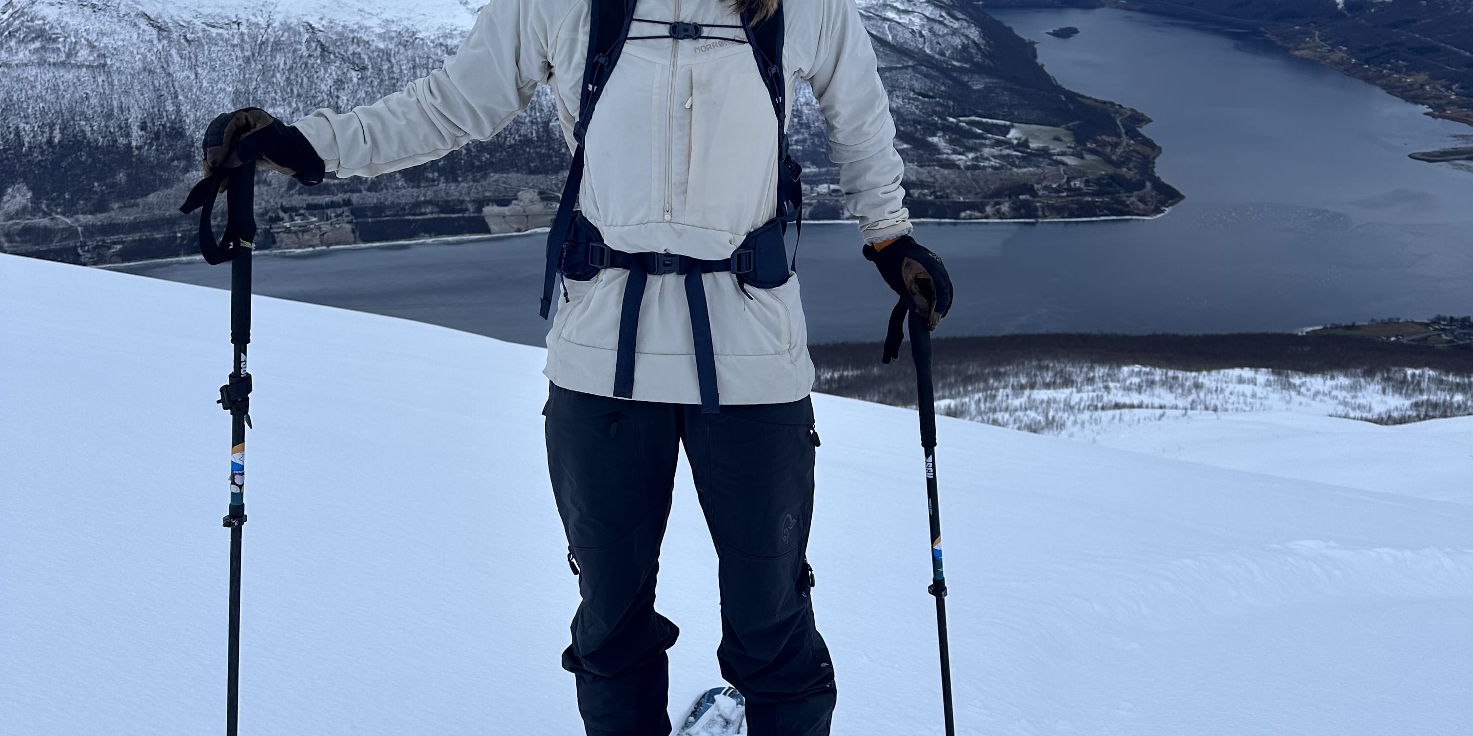 A smiling woman in a Norrøna jacket and hat skies in snowy mountains. The fjord is in the background.
