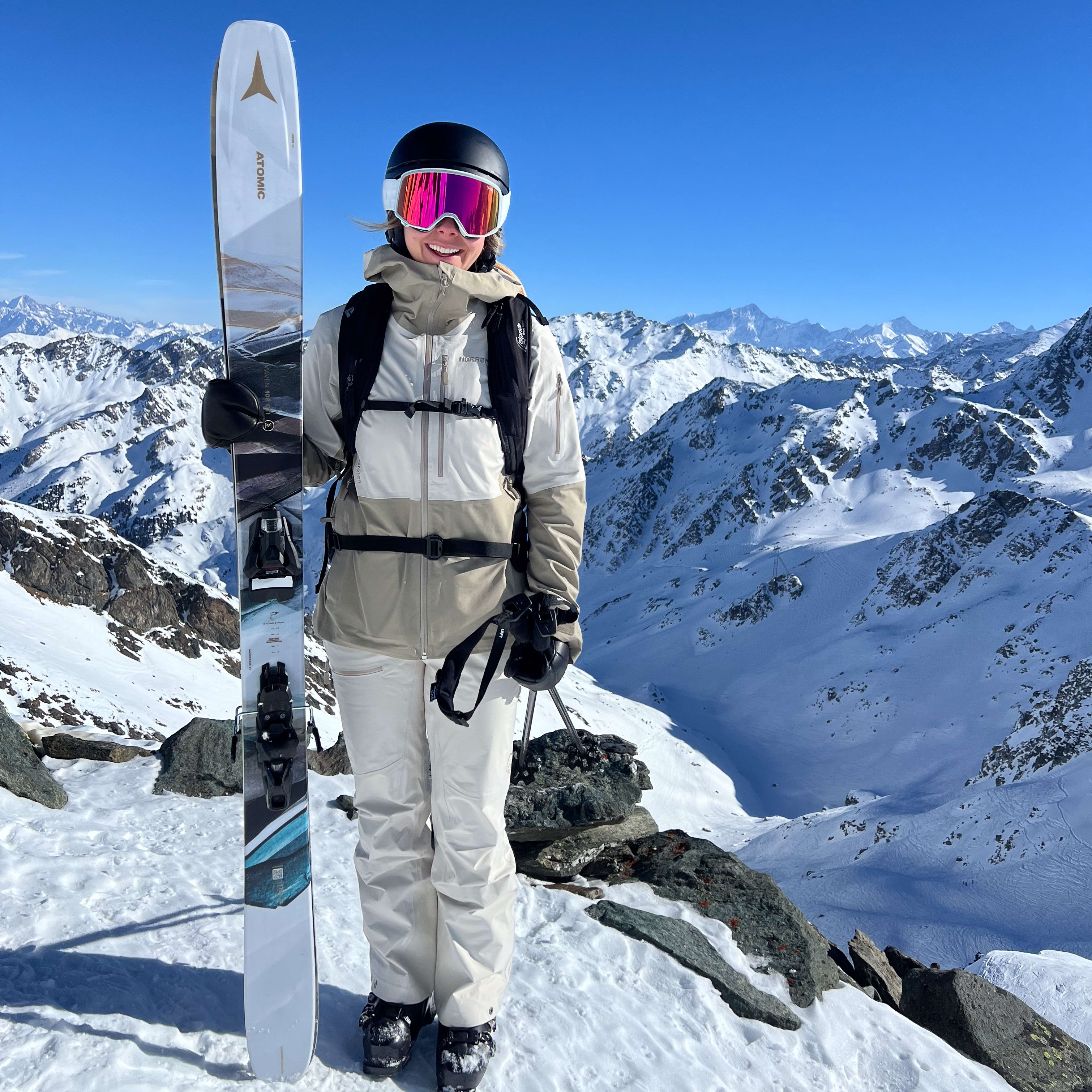 A woman wearing a Norrøna ski jacket and pants stands with a ski in front of a snowy mountain landscape. 
