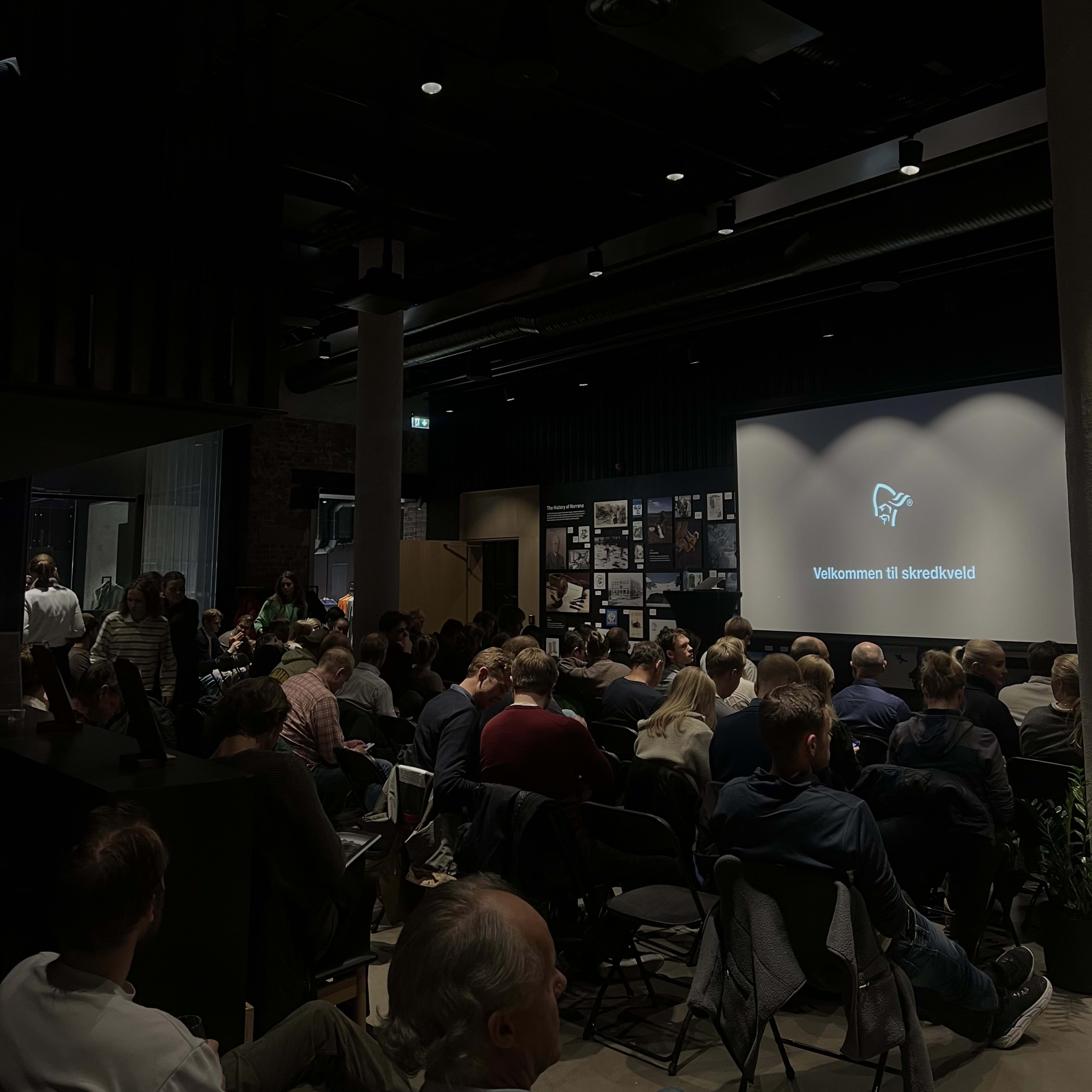 Audience in Norrøna clothing seated for a presentation showing "Velkommen til skredkveld" on screen.