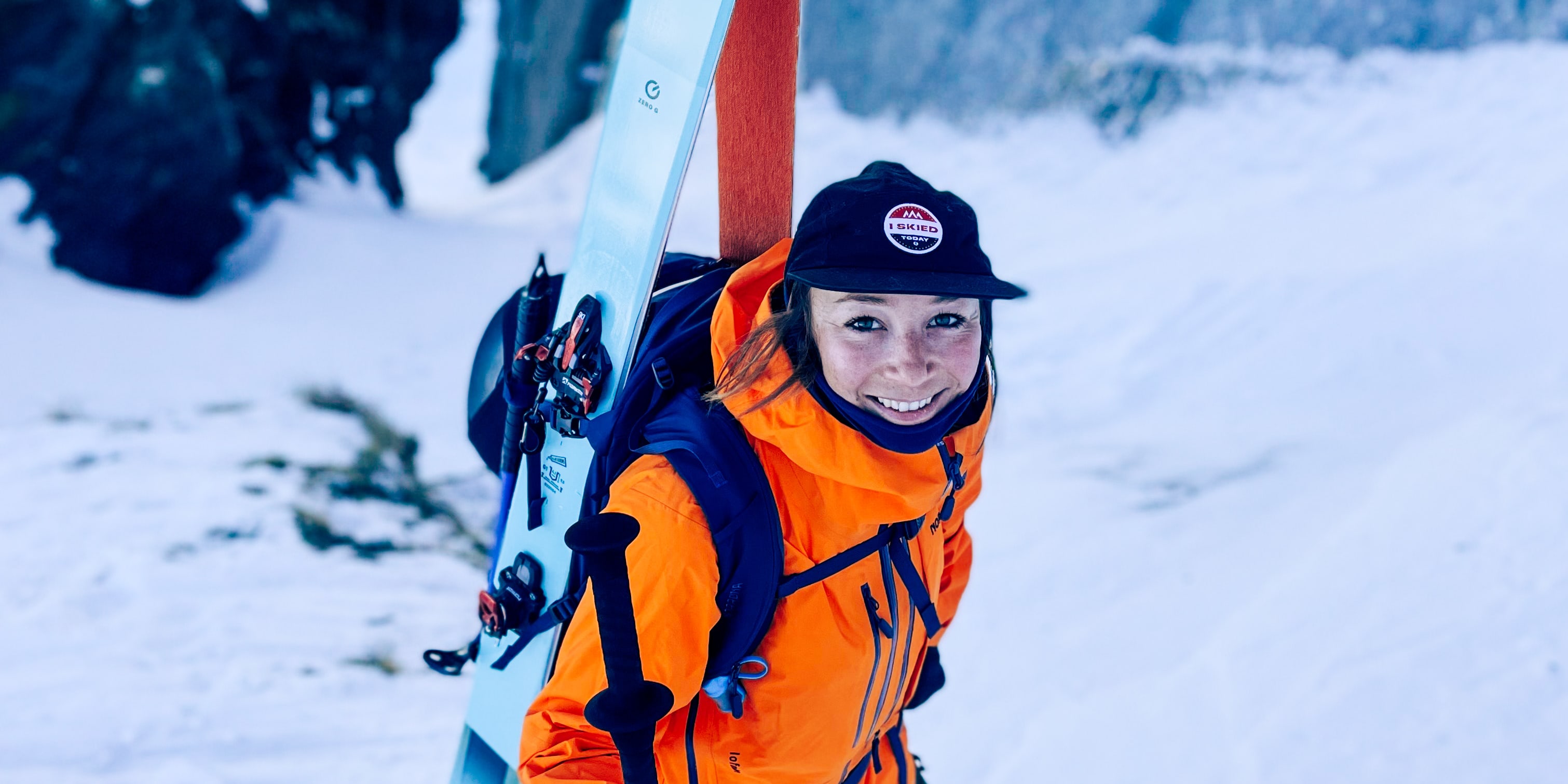 Smiling skier in snow. She's wearing a Norrøna orange jacket, carrying skis and poles. Rocks in background.