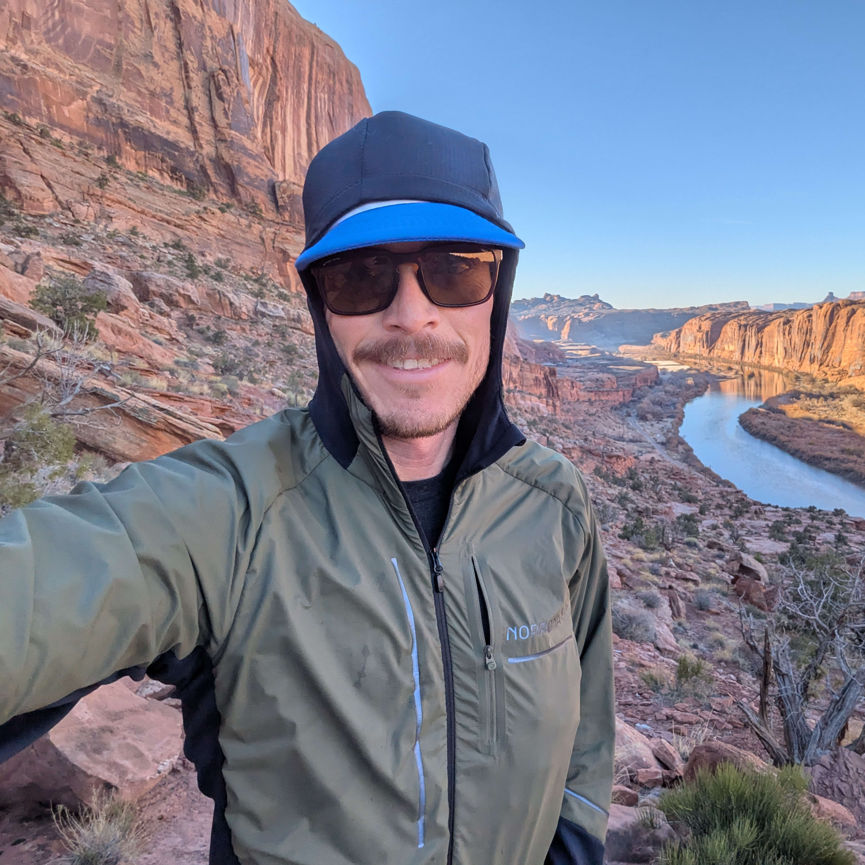 A man wearing a Norrøna jacket, sunglasses and a cap smiles while standing on a cliff overlooking a river canyon. 
