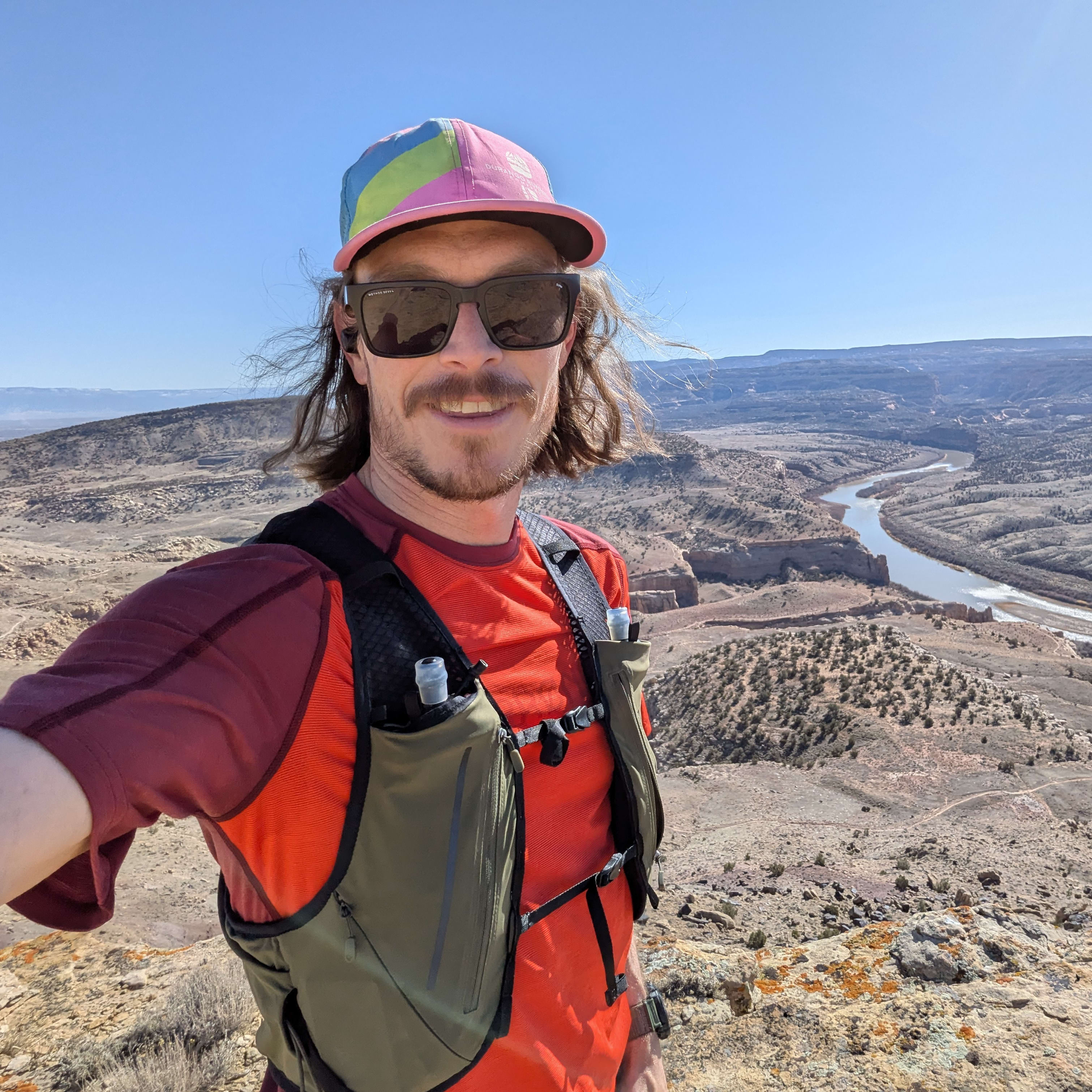 A person wearing sunglasses and a Norrøna running vest smiles while standing on a cliff overlooking a river winding through a desert landscape. 
