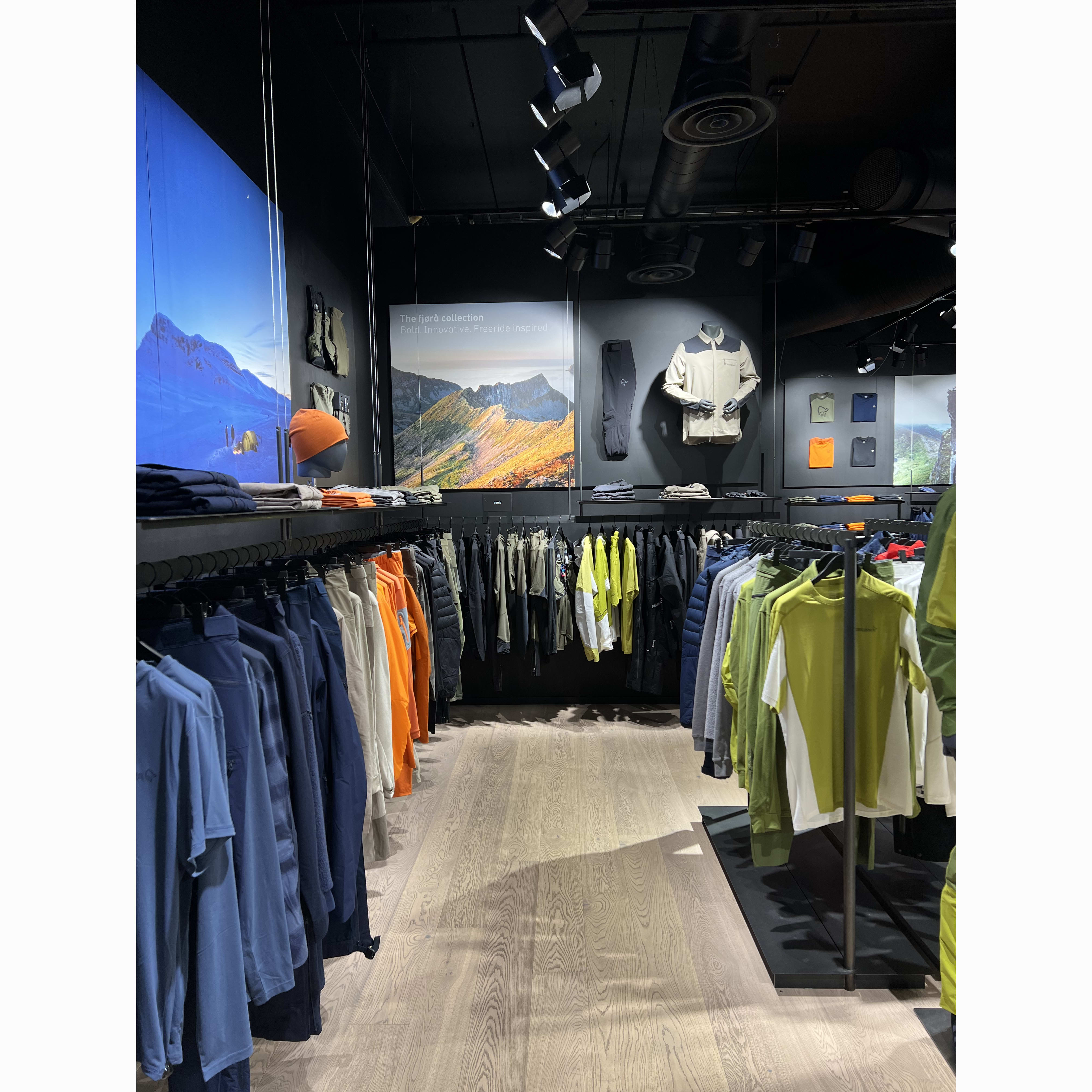 A view of a Norrøna clothing display in a store. Garments in blue, orange, and green hang on racks against a dark wall with mountainscape images.