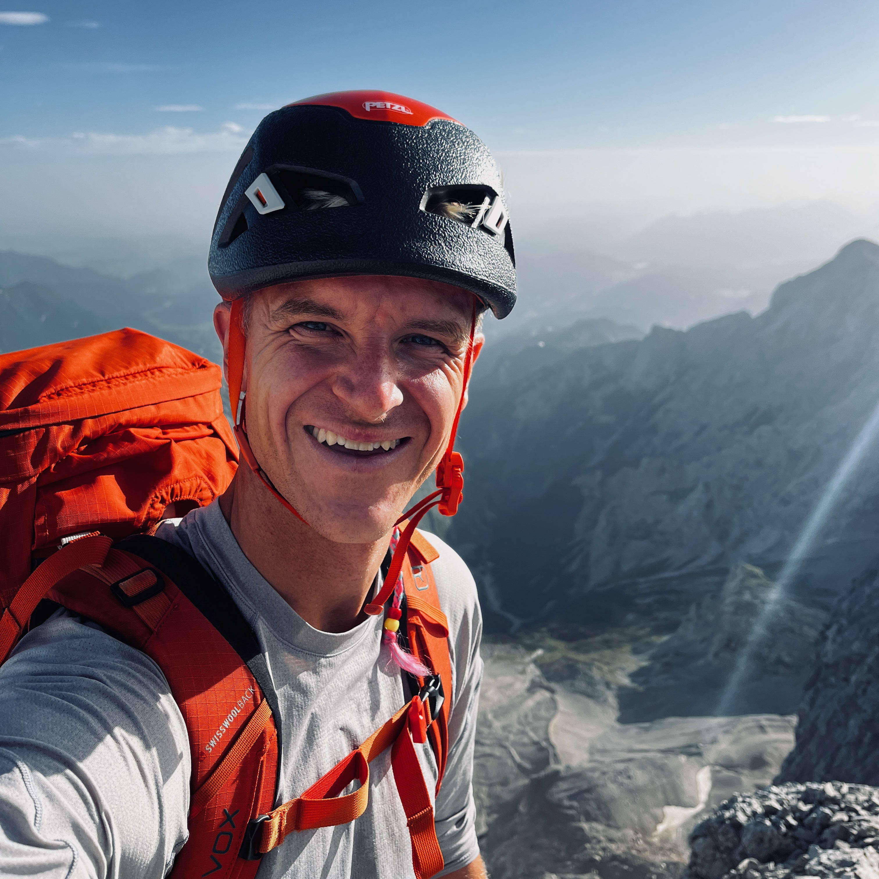 Smiling hiker with Petzl helmet and orange backpack, stands atop a mountain with hazy peaks in the background.
