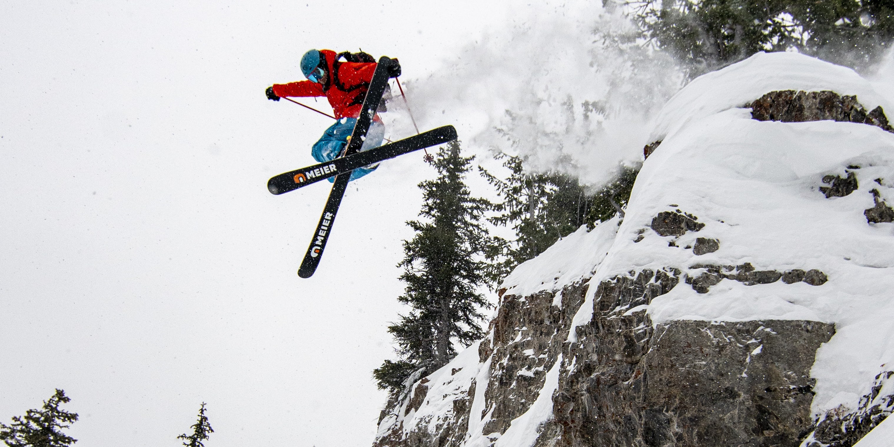 A skier in a red Norrøna jacket jumps off a snow-covered cliff with Meier skis against a snowy, forested backdrop.