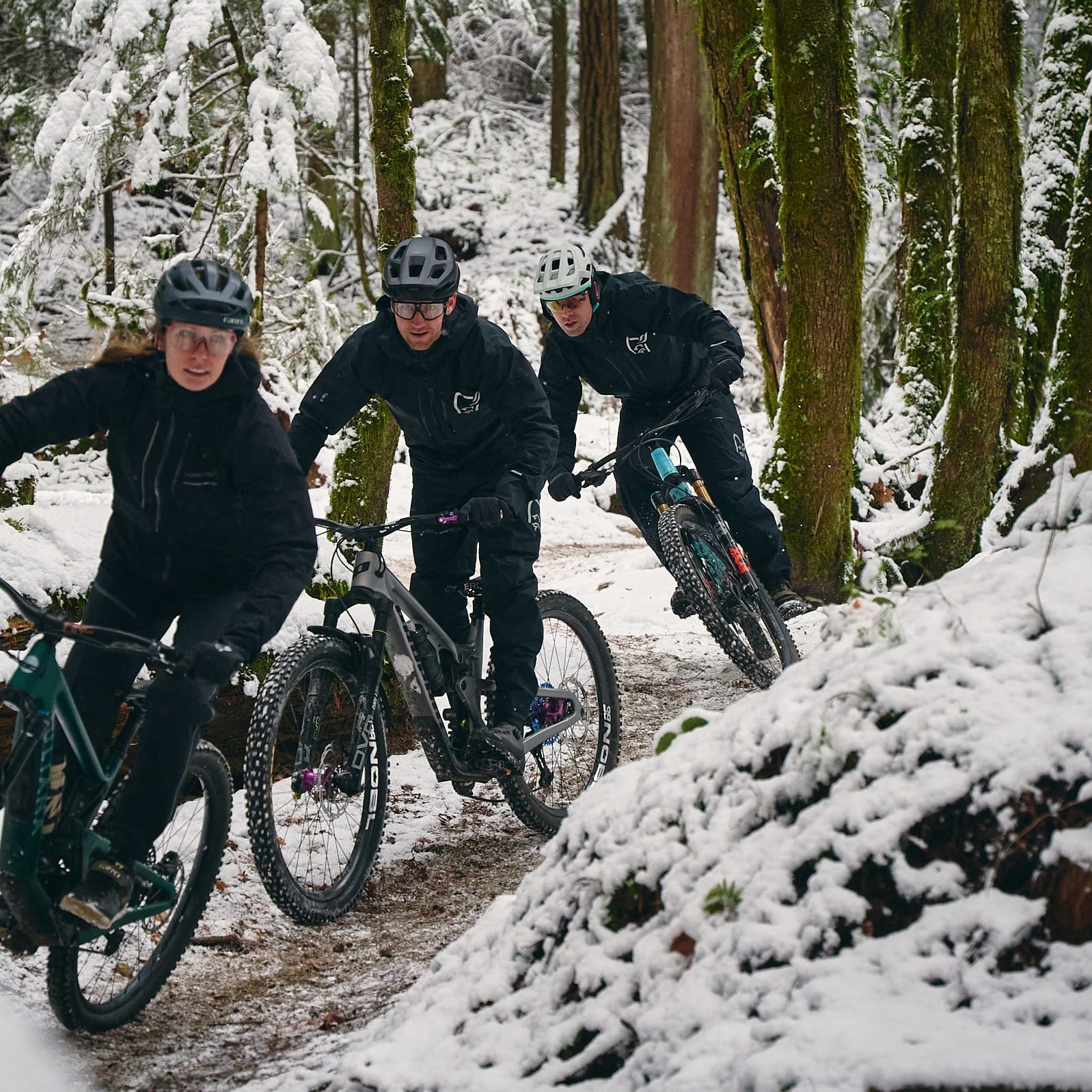 Three people wearing helmets and Norrøna gear ride mountain bikes through a snowy forest.  
