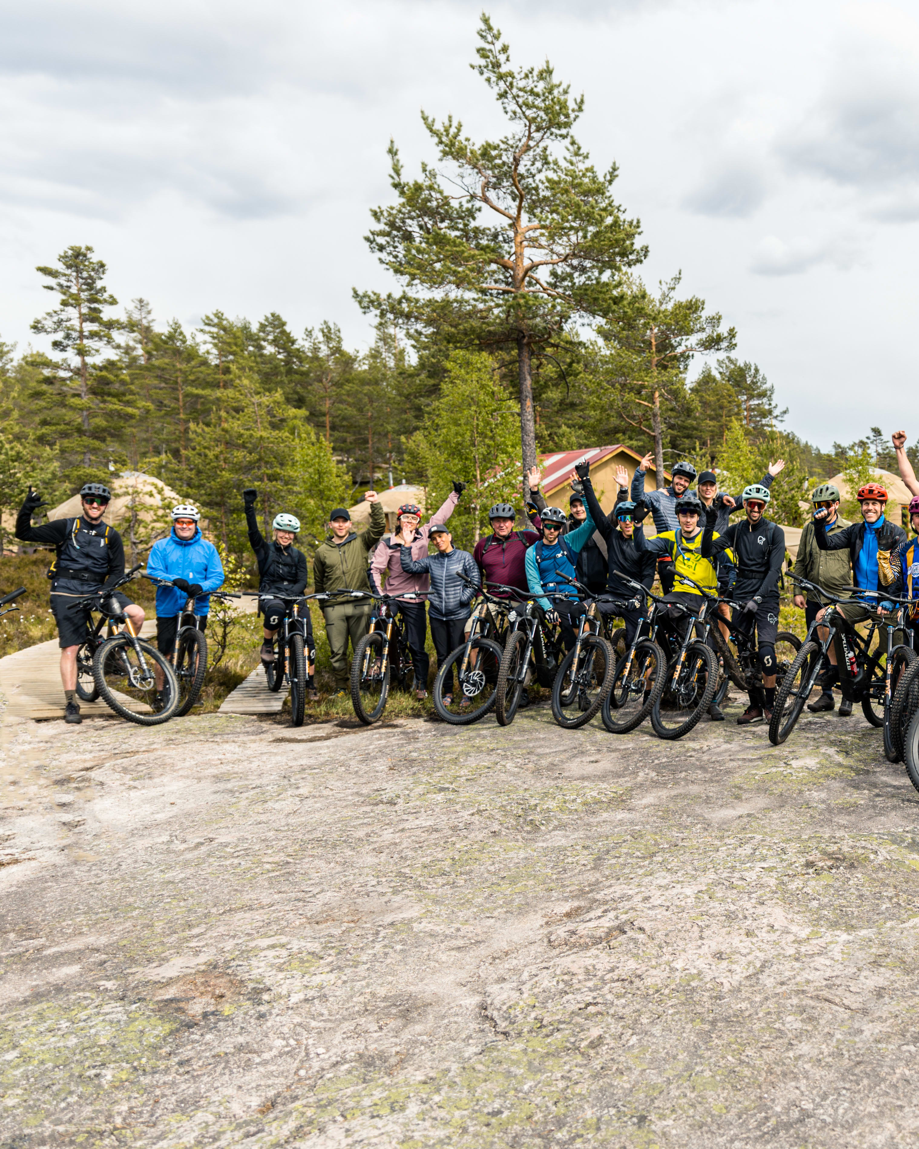 A group of mountain bikers, wearing helmets and Norrøna apparel, with their bikes, pose on rocky terrain with trees and a cloudy sky in the background.