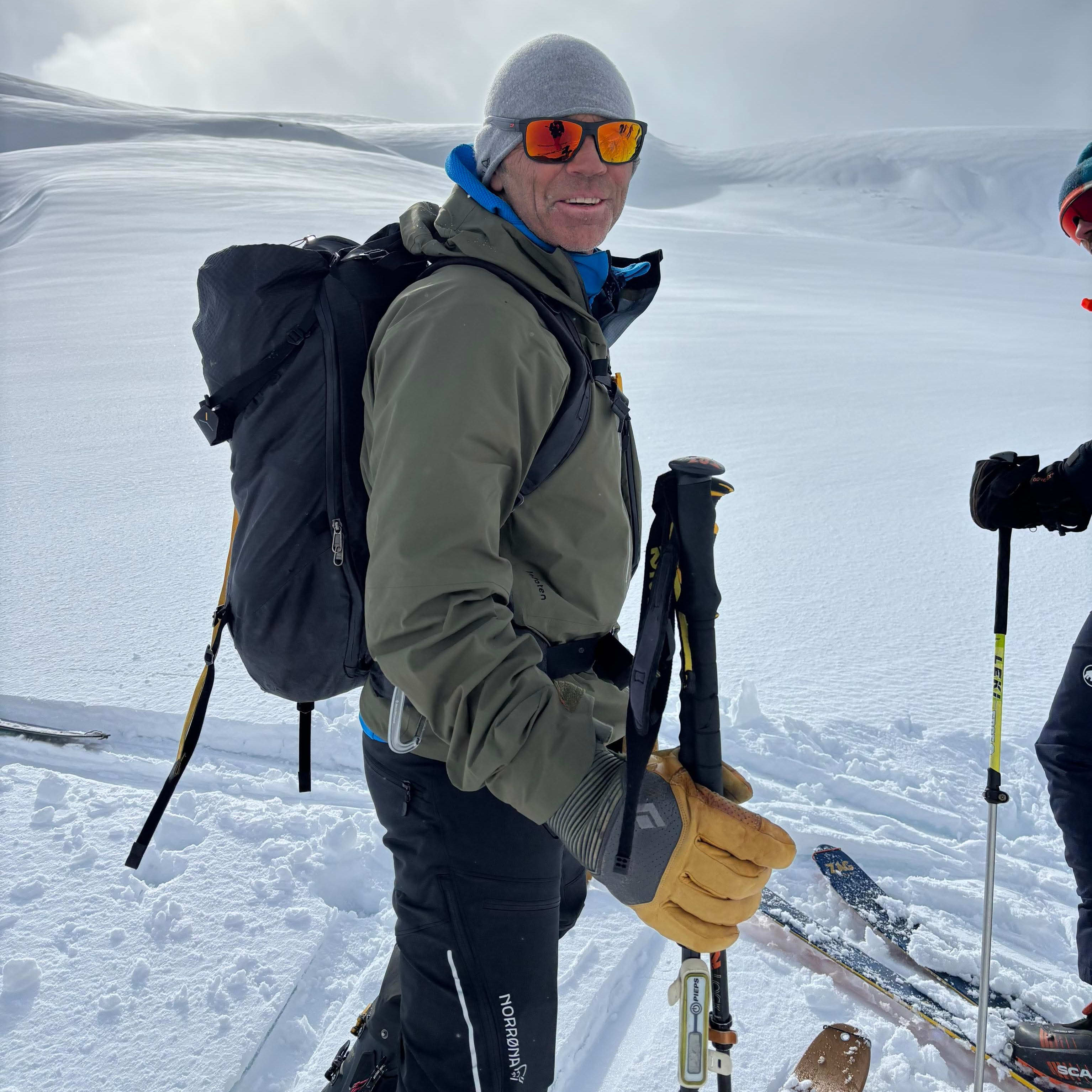 A man in Norrøna ski gear stands in a snowy landscape with ski poles and skis.  He is wearing a grey hat, sunglasses, and gloves. The sun shines brightly in the background. 
