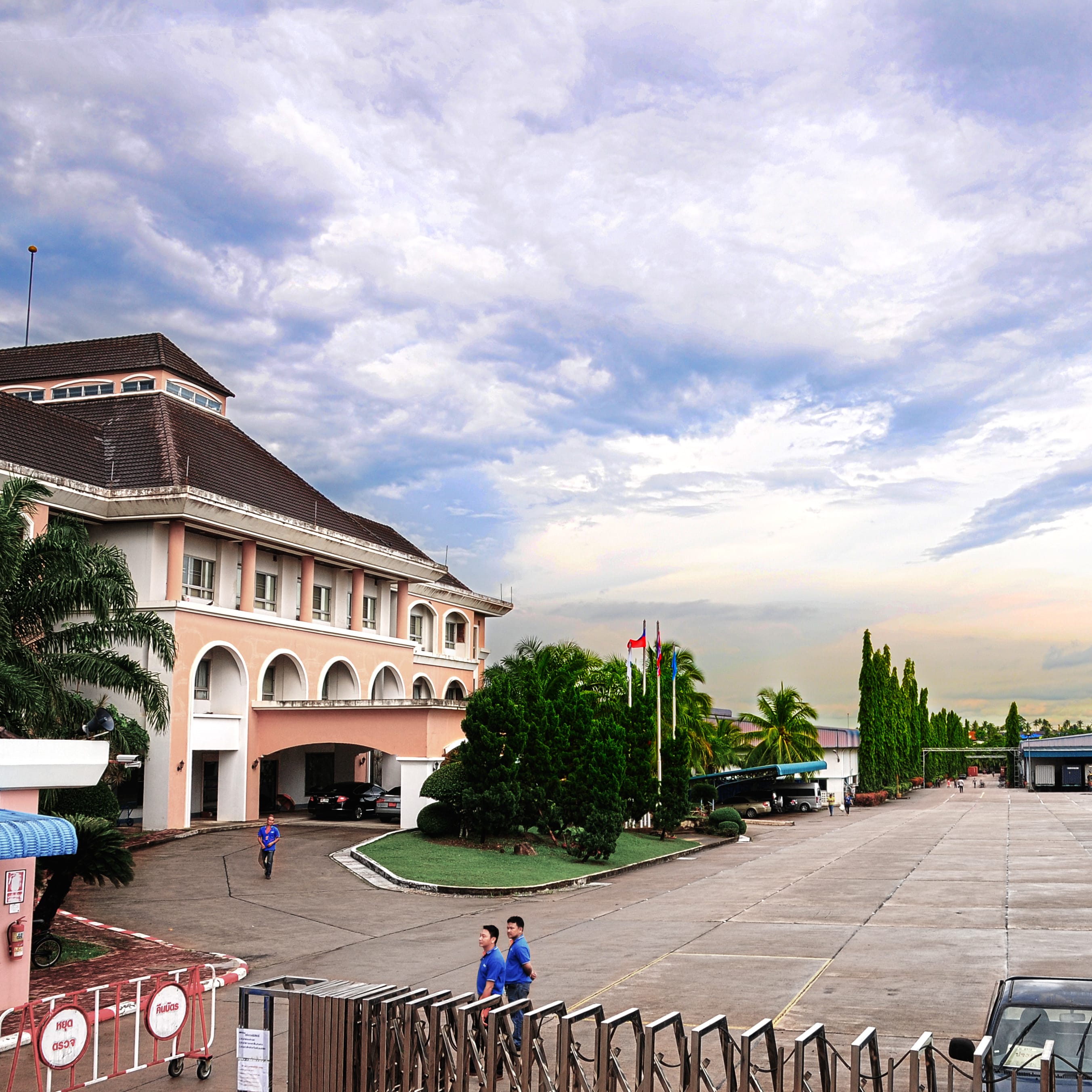A large industrial complex with an open parking lot, a large pink building in the foreground, and a large concrete building with a large metal roof in the background. There are two men in blue shirts walking on the paved parking lot, and there is a vehicle in the background with its trunk open. 
