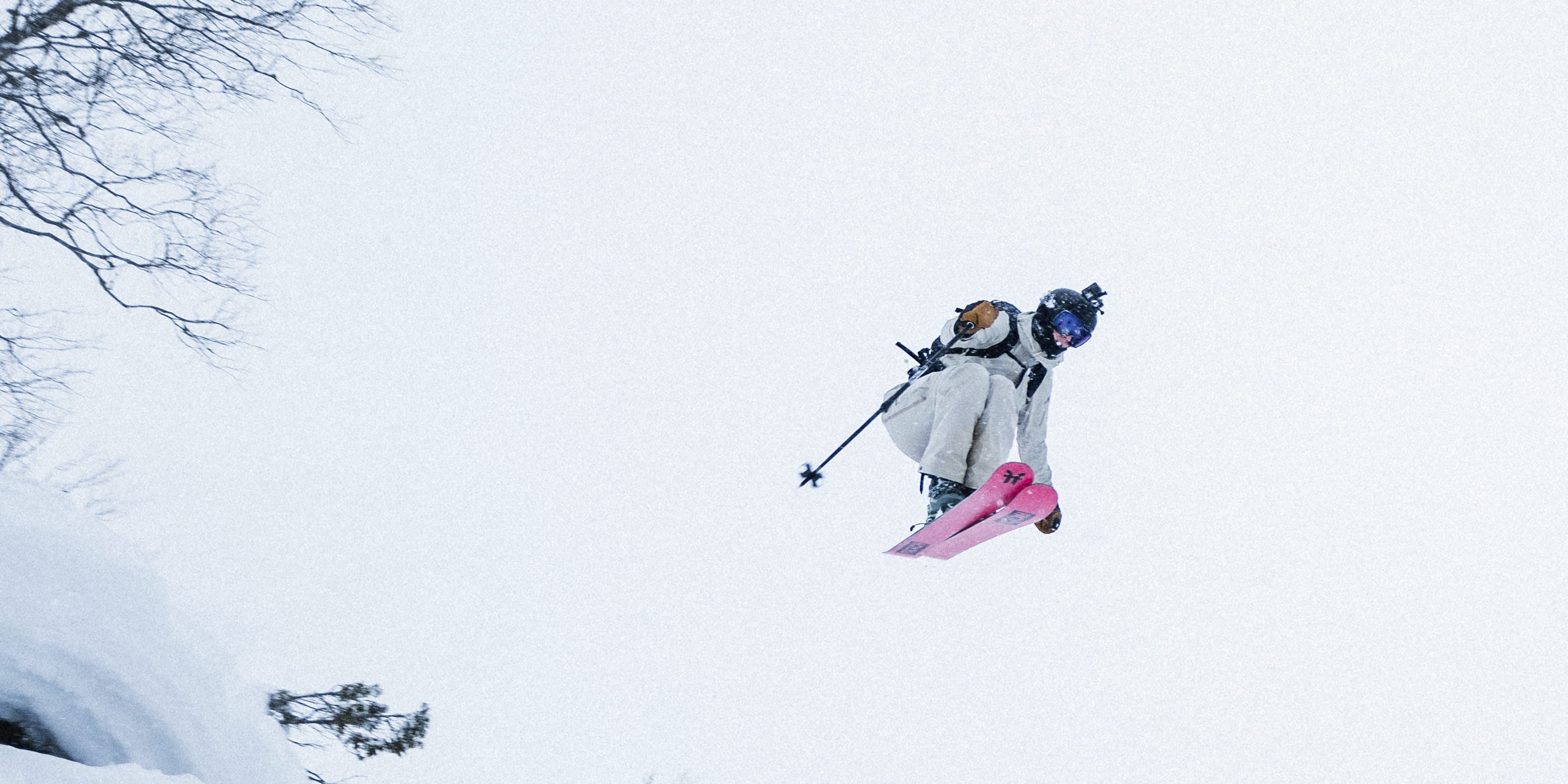 A skier wearing Norrøna clothing jumps off a snow-covered slope,  against a backdrop of a snowy mountain and trees. 
