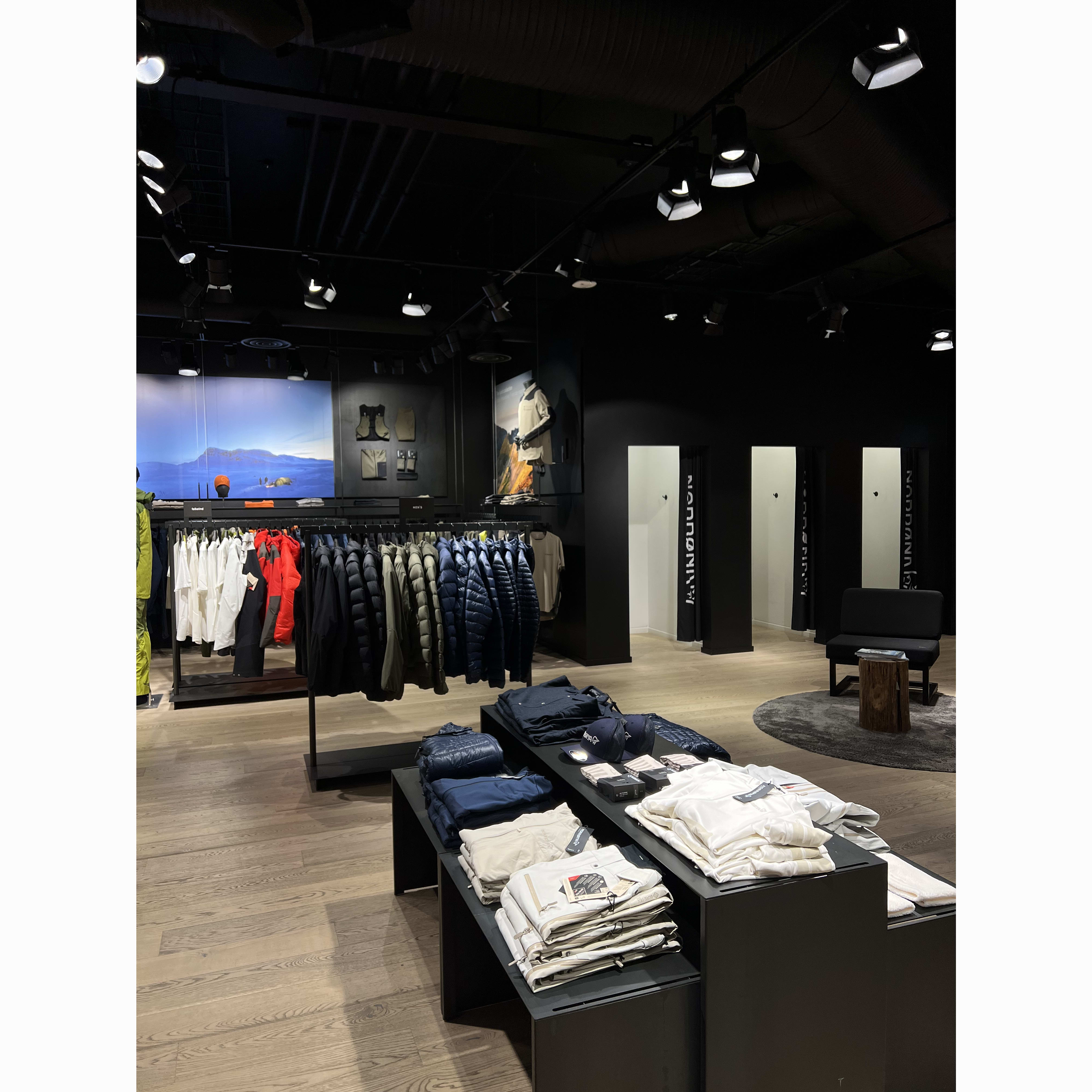Aisles in a Norrøna store display folded jackets and shirts on shelves and hangers. The store features a dark ceiling, bright lighting, and a wooden floor.
