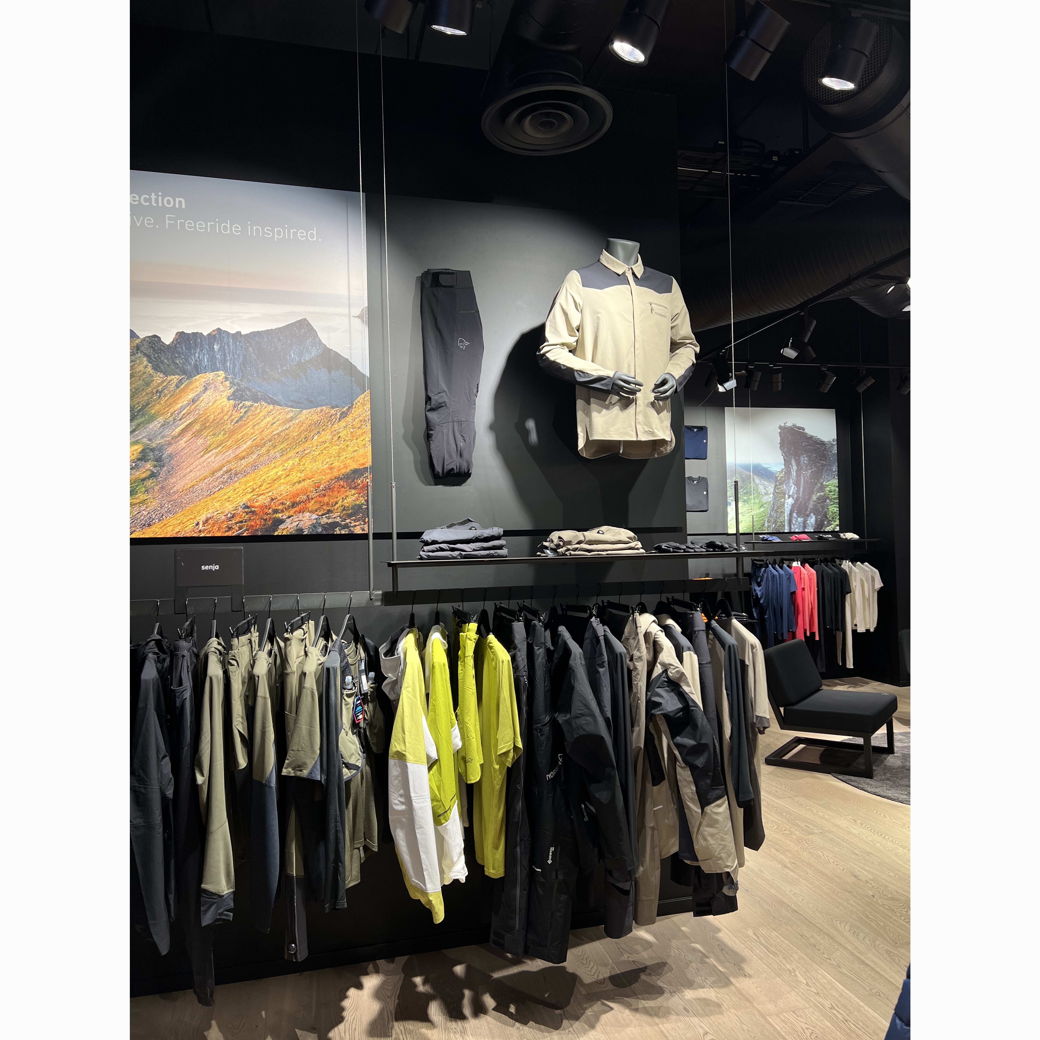 A store display features Norrøna clothing on black shelves and a wall, lit by spotlights. Jackets, pants, and colorful shirts are visible.