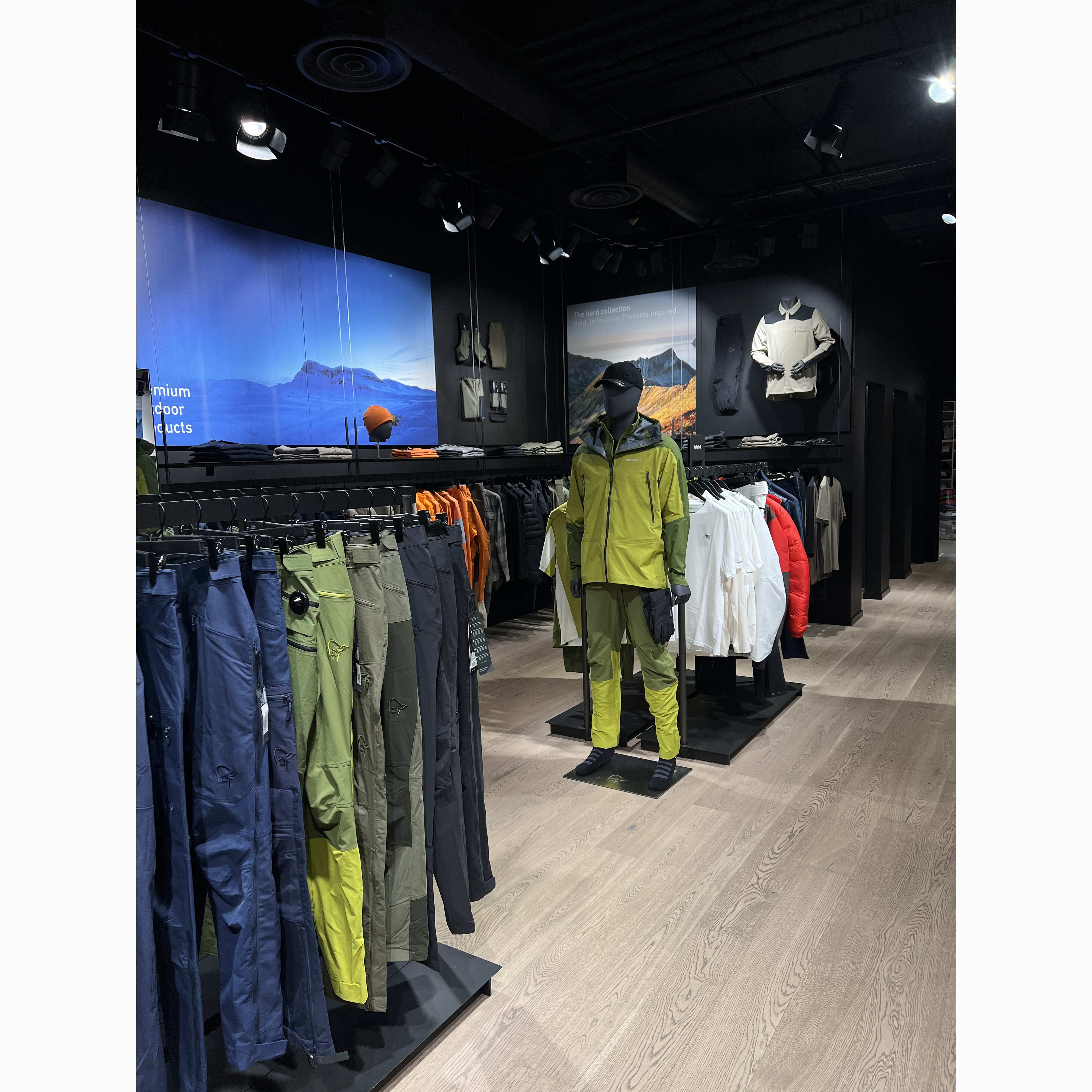 Norrøna store display: Mannequin wears green Norrøna jacket/pants. Racks of clothing flank display with mountain backdrop banners.