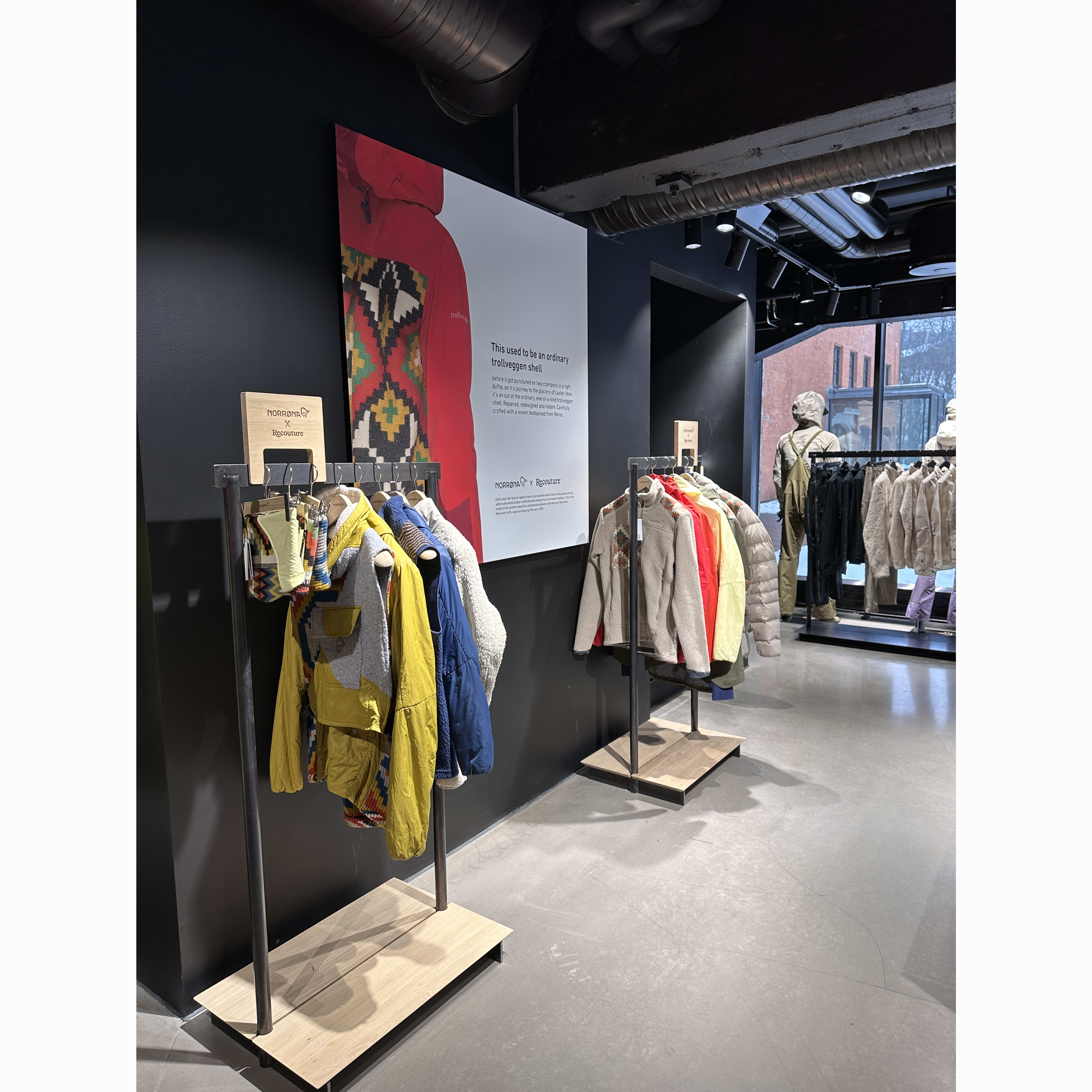 A retail display of Norrøna clothing in various colors stacked on metal racks against a black wall with a poster.
