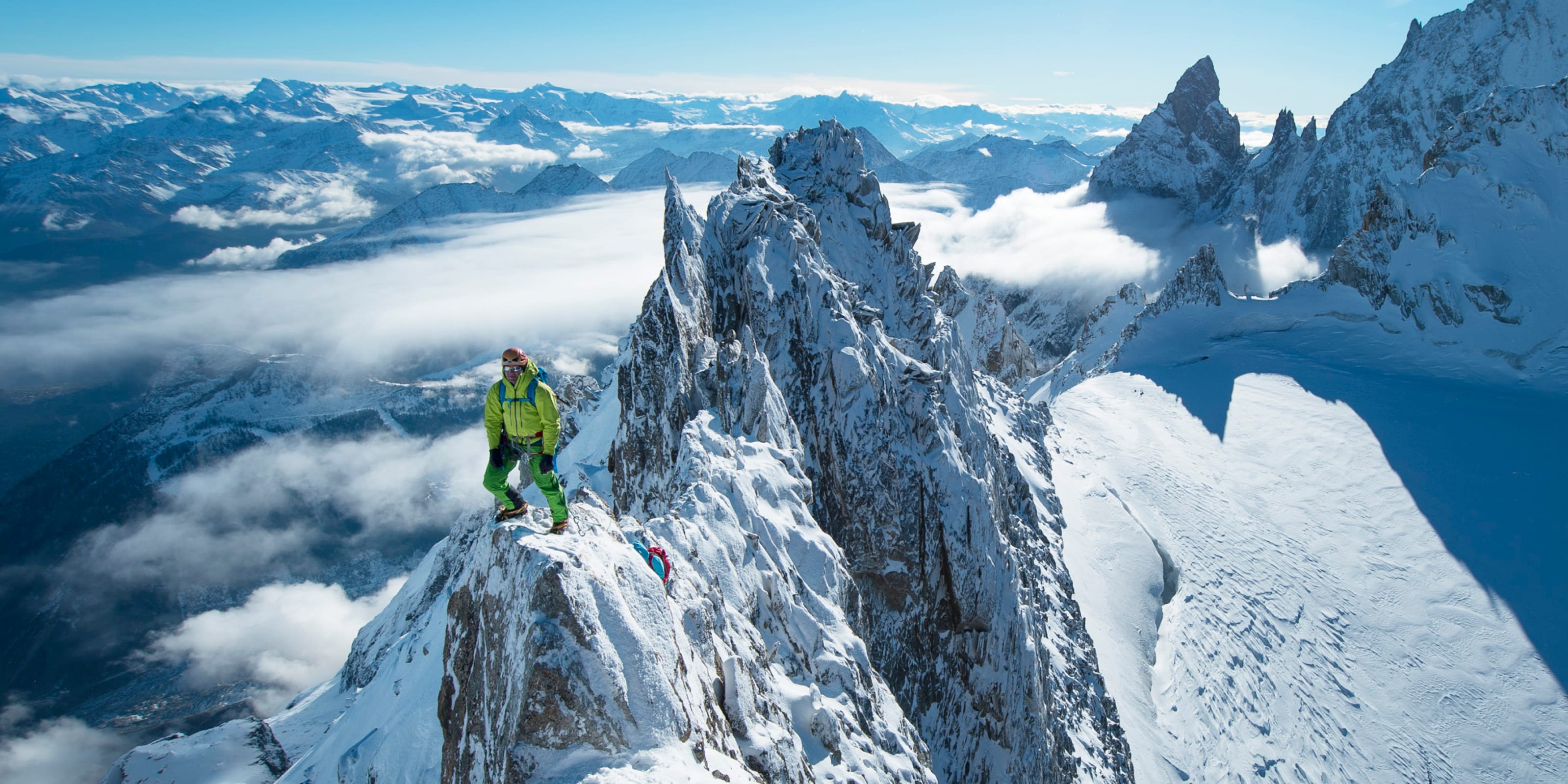 Two mountaineers ascend a snow-covered peak on a clear day, wearing Norrøna outerwear.  Mountains and clouds fill the background.

