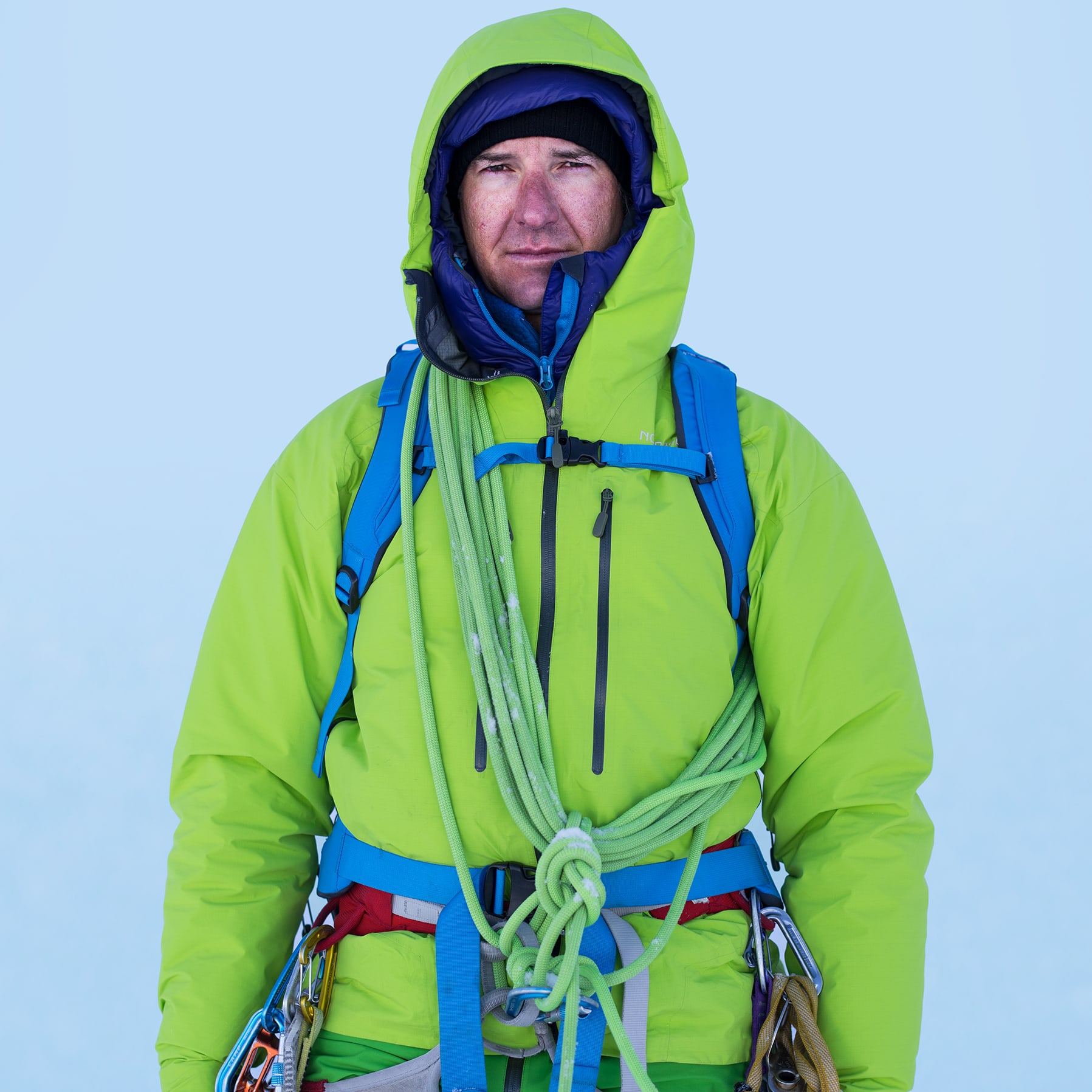 Mountaineer in Norrøna jacket, harness, and rope stands against a light blue snowy background.