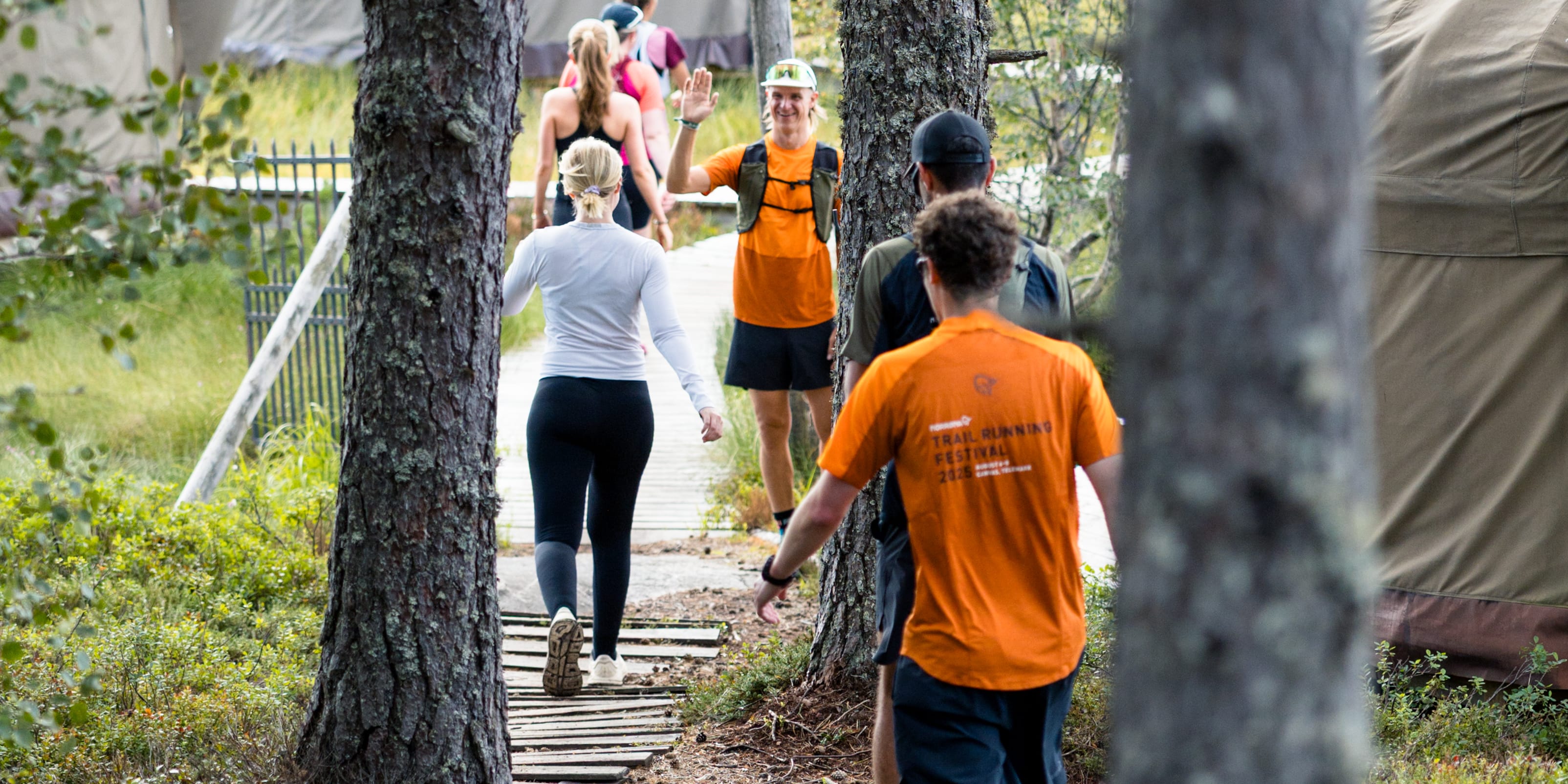 A group of people walks on a wooden path through a forest towards a tent encampment. One man waves wearing a Norrøna shirt.
