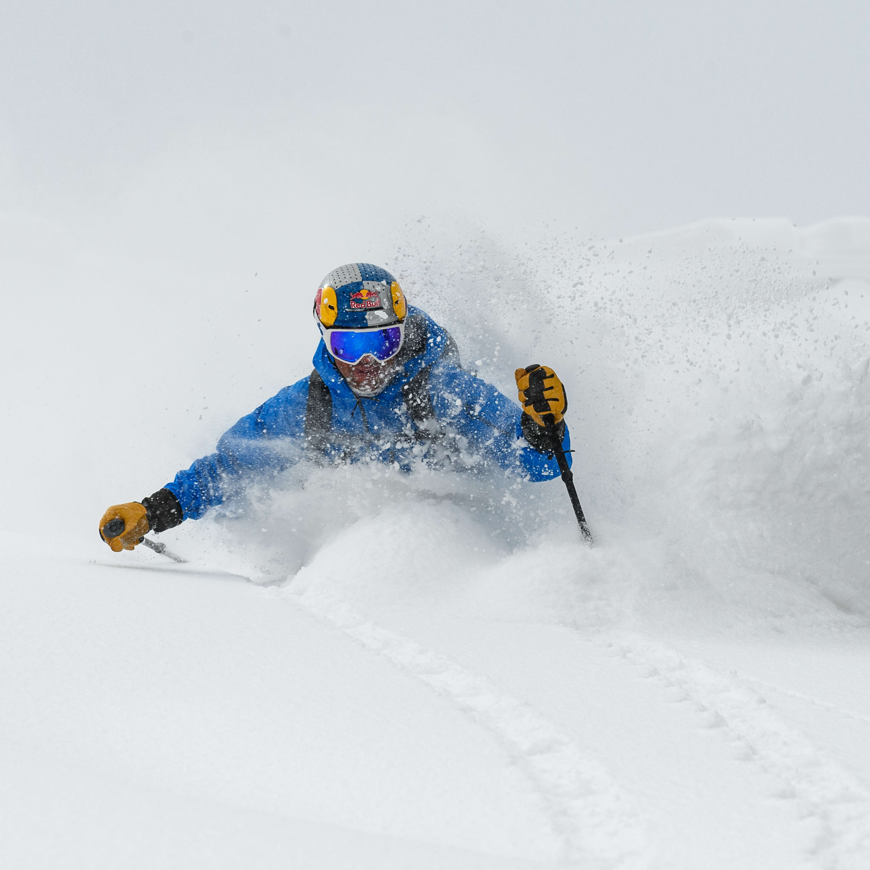 A skier in a blue Norrøna jacket carves through deep powder, kicking up a cloud of snow. They wear a Red Bull helmet and goggles.