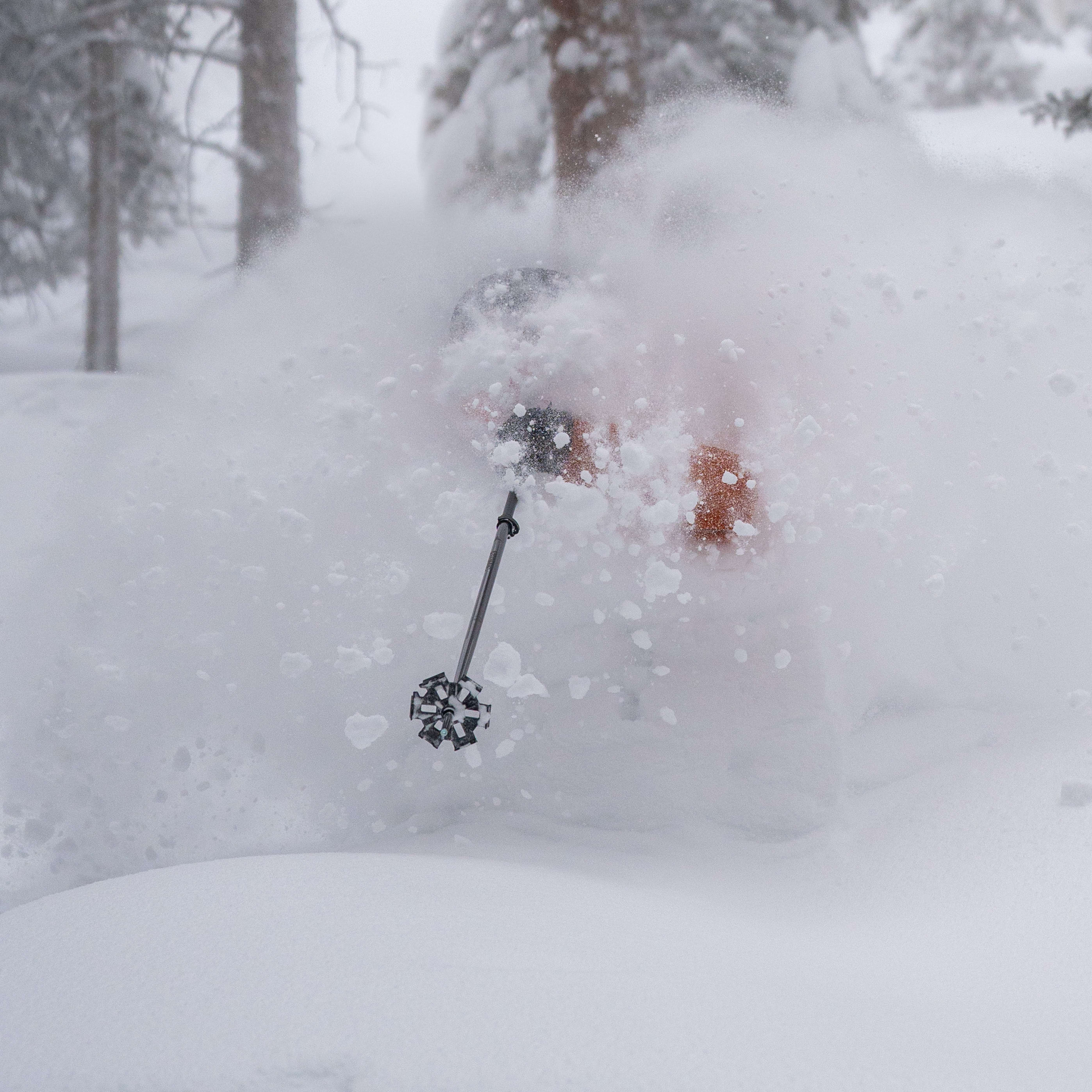 A skier in a Norrøna jacket and pants skis through deep powder snow. The skier's face and body are obscured by snow. The tips of the skier's ski poles are visible.