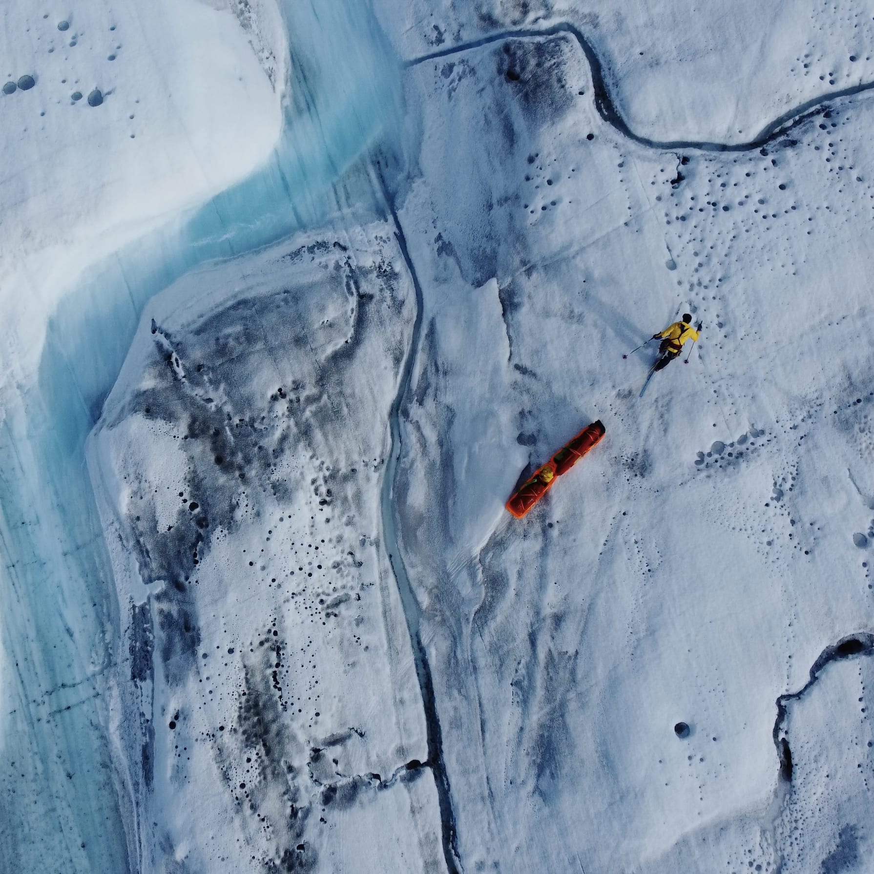 An aerial view of a glacier with a person in a yellow Norrøna jacket and orange Norrøna rescue equipment.  The glacier is covered in snow and ice, with a blue crevasse cutting through the center of the image.  The person is using a rescue sled. 
